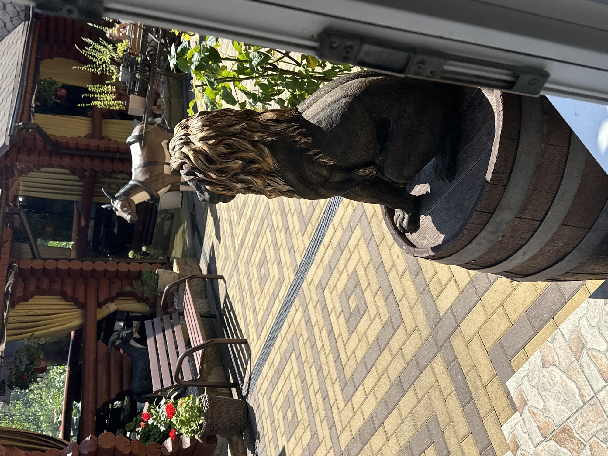 A sunny outdoor scene in Krasnaya Polyana, Russia, features a statue of a lion sitting on a wooden barrel in the foreground. The lion statue is dark in color with a mane of light brown and gold. Behind the statue, a paved courtyard with a geometric pattern of yellow and gray bricks is visible. To the left, a dark brown bench sits near a potted plant with red flowers. Further in the background, a wooden structure with a thatched roof is partially obscured by yellow curtains. A dark-colored goat statue stands near the bench, and a brown and white horse-drawn cart is situated further back. The environment appears to be a landscaped area, possibly a garden or a public space in Krasnaya Polyana.
