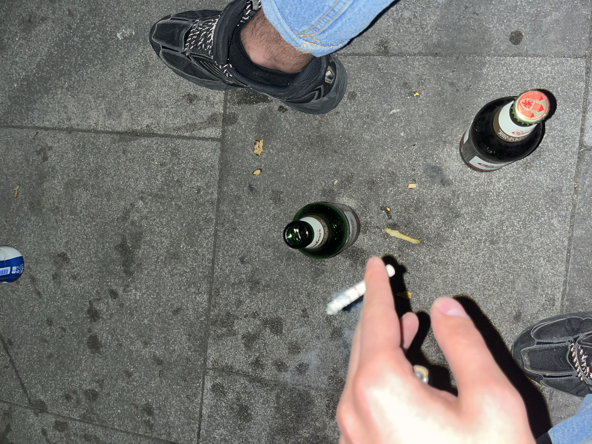 The image presents an overhead view of a street scene at night, likely in an urban area like Sofia, Bulgaria. The ground is paved with dark grey, textured slabs, showing some scuffs and scattered debris.
Two individuals are partially visible in the frame. In the upper left quadrant, a person's left leg is shown, clad in blue jeans and a black athletic shoe with reflective details. The ankle is bare and hairy. In the bottom right corner, another person's right leg is partially visible, also wearing blue jeans and a dark sneaker.
Several objects are placed on the ground. A dark brown beer bottle is prominently featured on the right side, with a distinct red cap bearing a red leaf symbol and the brand name "COORS" partially visible on its label, indicating it's likely Coors Light. Another green glass bottle lies in the center, near the first bottle. In the lower center, a hand holds a lit cigarette, with a visible wisp of smoke rising from it. Scattered around the bottles and on the ground are several small, yellowish pieces of food, resembling french fries. A small blue and white object is also partially visible on the far left edge of the frame.
The lighting suggests it is evening or night, with visible reflections on the shoes and limited ambient light. The scene depicts a casual outdoor gathering or break, involving drinking, smoking, and possibly snacking.