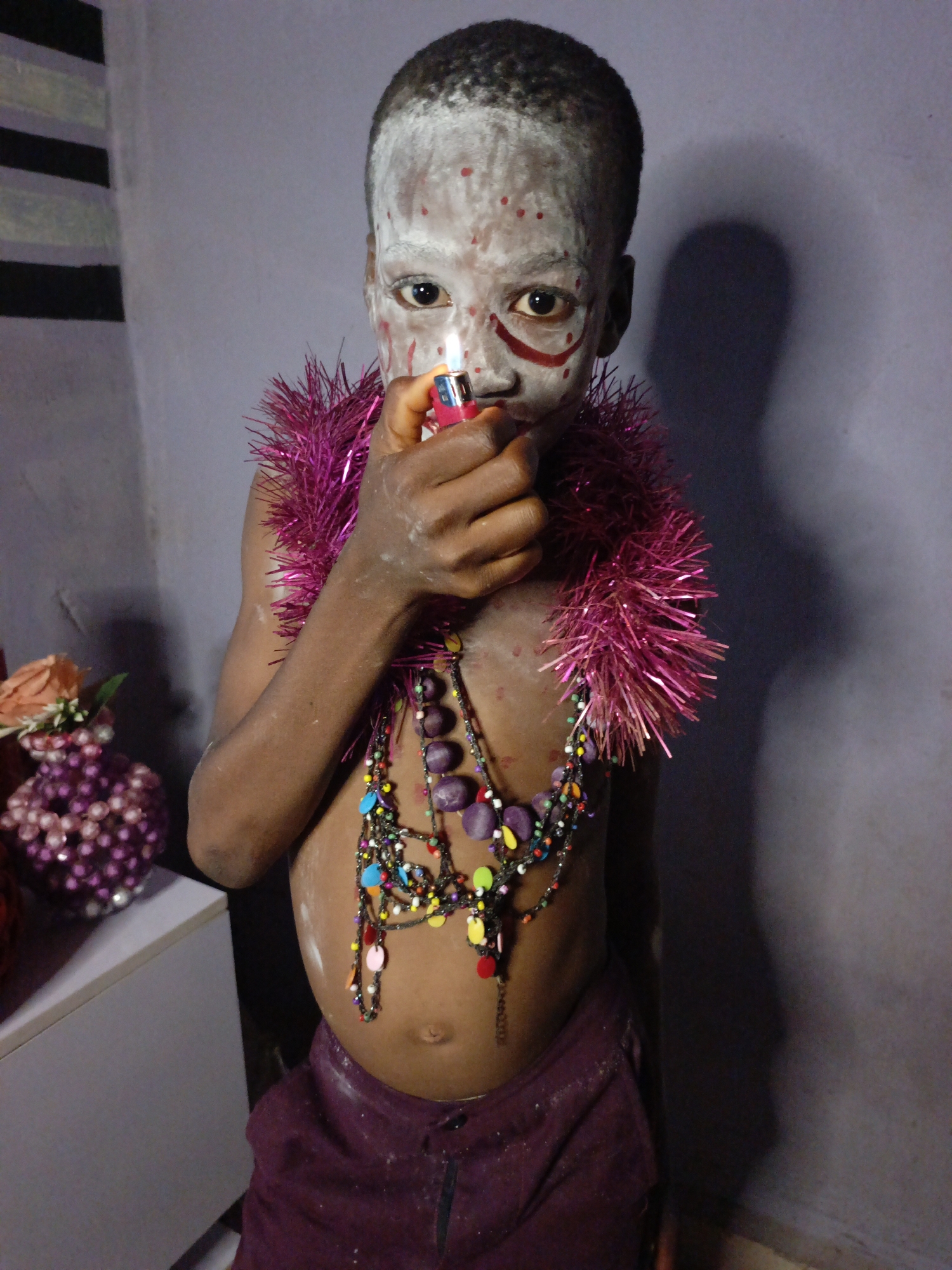 An indoor portrait captures a young child, likely male, looking directly at the camera. The child's face and forehead are painted white with numerous small red dots and two bold, red, curved lines under each eye. The child's dark hair is closely cropped. Around the child's neck and draped over the shoulders is a vibrant, metallic pink tinsel boa or garland. Multiple beaded necklaces are worn over the bare chest and stomach, featuring a mix of large, smooth purple beads and smaller, multi-colored beads in shades of yellow, blue, red, orange, and black.
The child holds a pink and silver disposable lighter, which is actively lit, producing a small flame near the lower part of the face. The hands and arms show smudges of white paint or powder. The child is wearing dark purple or maroon shorts/pants.
The background is a solid, light purple-grey wall. To the far left, a section of the wall features vertical black and white stripes. On a light-colored surface to the left, a decorative object made of purple beaded spheres, possibly a vase, is visible, with a light-colored flower or embellishment beside it. A dark shadow is cast on the wall to the right of the child, suggesting indoor lighting conditions. No text is visible in the image. The child's expression is direct and focused.