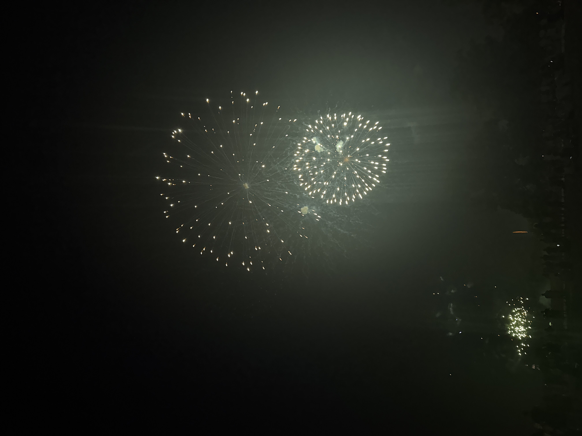 The image depicts a nighttime fireworks display against a dark sky. Two large, circular fireworks with bright white bursts are prominently featured in the upper half of the frame, with one slightly above and to the left of the other. Smaller, scattered fireworks are visible in the lower left and lower center portions of the image, some appearing as clusters of light. A faint green haze or smoke permeates the lower half of the scene, obscuring the details of the ground. Silhouetted figures of a crowd are visible along the bottom of the frame, suggesting an outdoor event. The event is taking place in Lille, France.