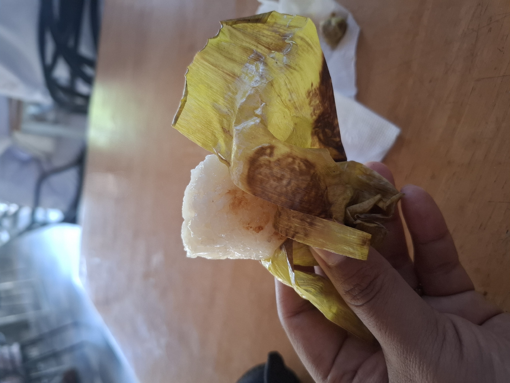 A hand is holding a small, white, cylindrical food item wrapped in a green, dried leaf. The food item appears to be sticky rice. The leaf is textured and partially covers the food. The background shows a wooden table and a blurred interior scene with chairs and people. The scene is likely in Keuniree, Indonesia, where this type of food is common.