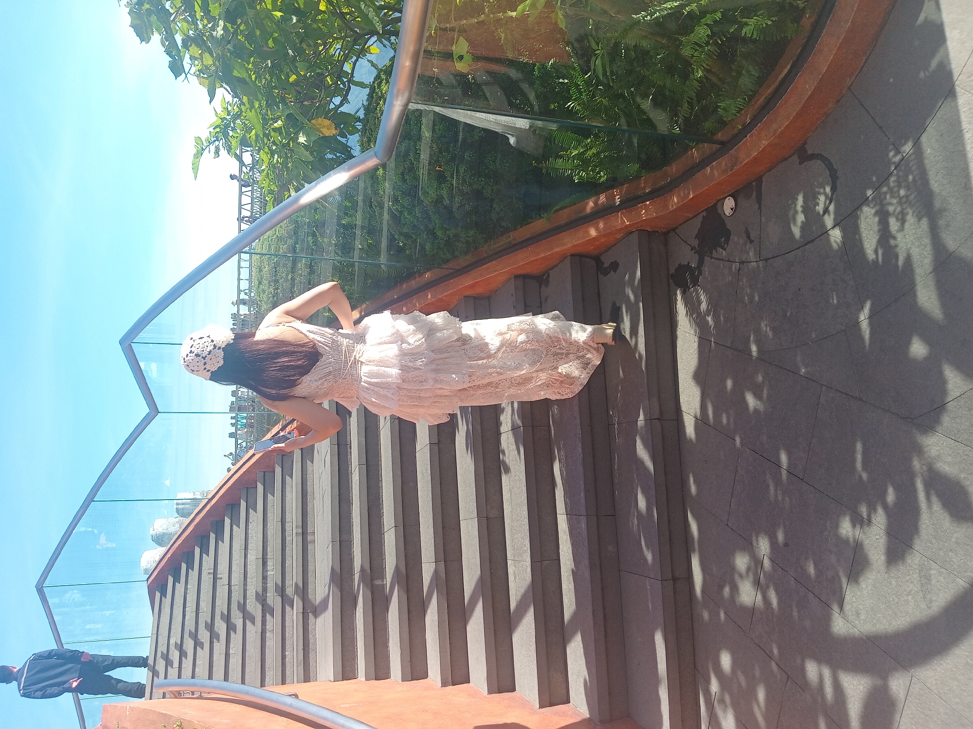 A woman in a light-colored, tiered dress and a straw hat is walking up a wide, outdoor staircase. She is holding a smartphone in her right hand. To her left, further up the stairs and facing away from the camera, is a man in dark clothing. The staircase has a modern design with concrete steps and orange-red concrete sides. A glass railing with a silver-colored metal handrail runs along the right side of the stairs, offering a view of lush greenery and foliage below. The scene is outdoors under a bright blue sky with scattered clouds. The image appears to be taken in Đà Nẵng, Vietnam, possibly at a scenic viewpoint or attraction. The lighting suggests it is daytime.