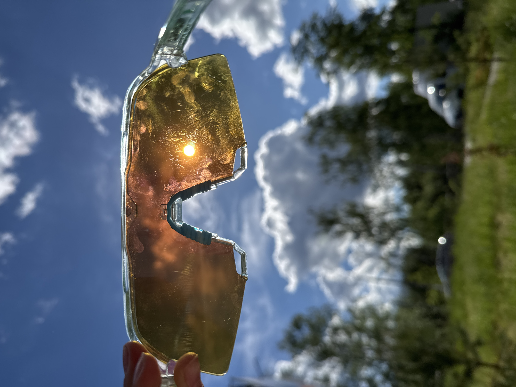 The image shows a pair of yellow-tinted sunglasses being held up against a sky with blue patches and white clouds. Through the lenses, the sun is visible as a bright spot. Behind the sunglasses, in the background, are blurred trees and a hint of a vehicle. It is not possible to ascertain the exact location in the city of Chaikovsky, Russia, from the given information.