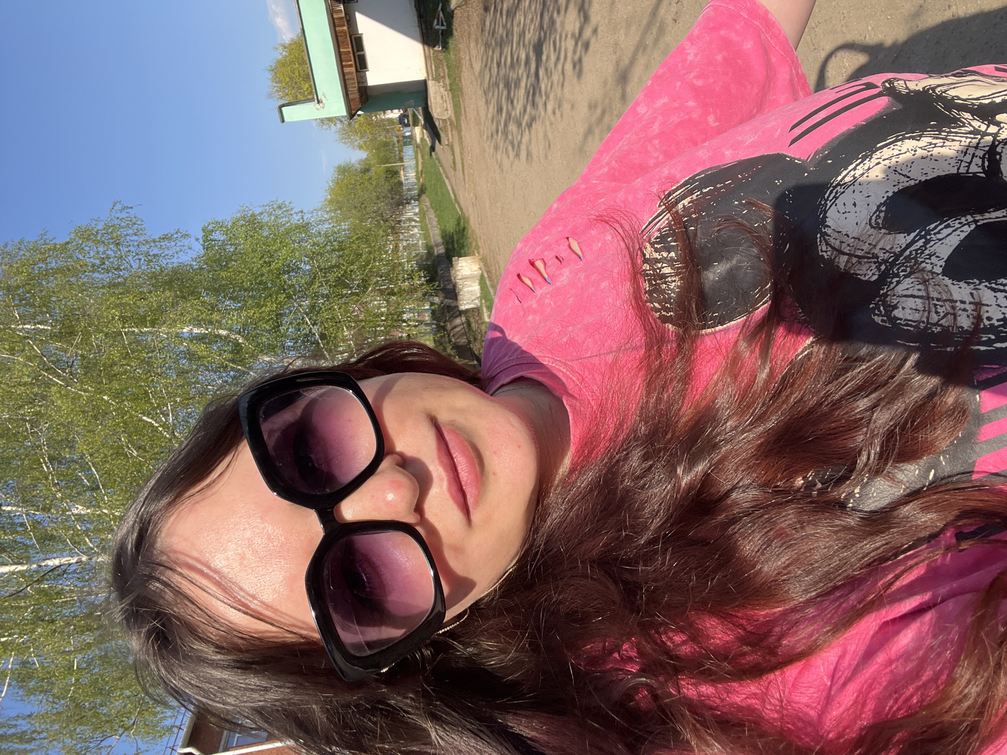 A woman with long brown hair, wearing large black sunglasses and a pink t-shirt, is centered in the foreground. The background includes green trees, a light blue sky, and a building with green trim. The location is in Noviy, Russia, as implied by the description. The woman is likely outdoors on a sunny day.