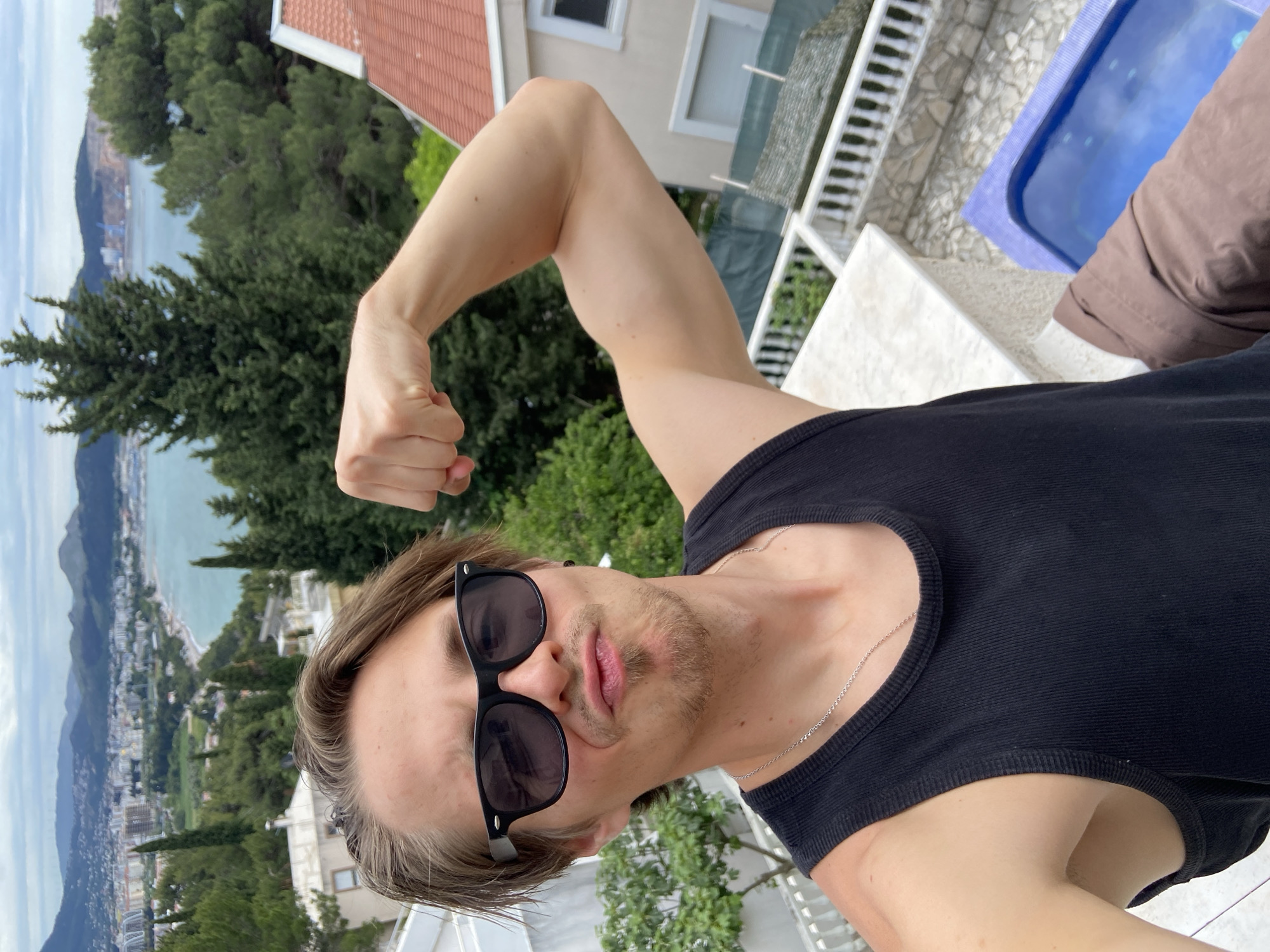 A fair-skinned person wearing a black tank top and sunglasses poses for a selfie, flexing one arm towards the camera. The person has short blonde hair, facial hair, and a thin silver necklace. Behind the person is a cityscape and coastline of Šušanj, Montenegro. Trees and buildings are visible, along with a body of water in the distance. A white railing and a small pool are visible near the subject.