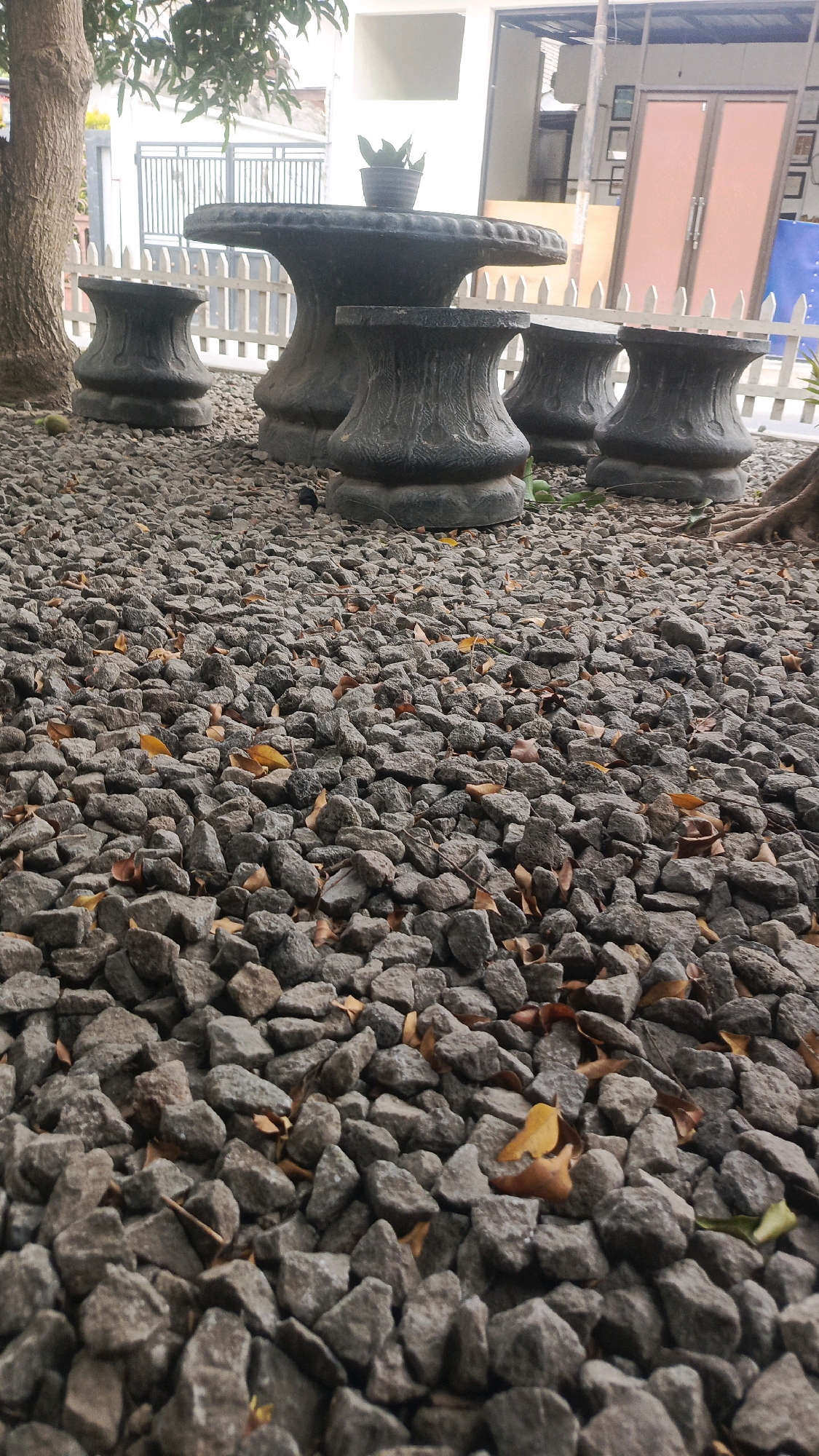 The image shows a stone table and four stone stools on a gravel-covered ground in front of a building with orange doors. There are some fallen leaves scattered on the ground and a small potted plant on the table. A tree trunk is visible on the left side of the image. The background shows a white picket fence and a building with windows and doors. The lighting suggests it is daytime. There are no people visible in the image.