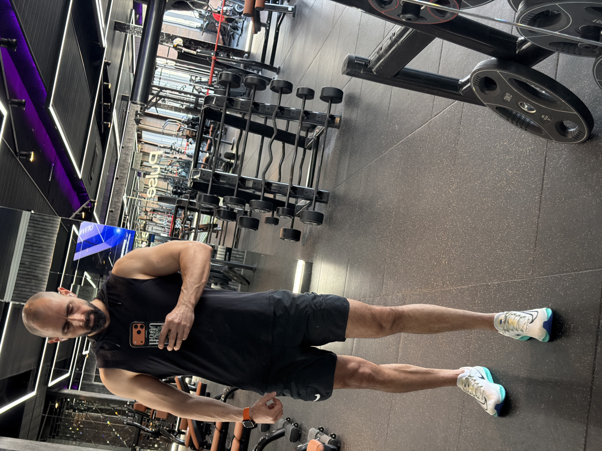 A man stands in a gym. He is bald with a beard, wearing a black sleeveless shirt, black shorts, and running shoes. He is holding a phone. The gym has various weightlifting equipment, including dumbbells and weight plates. The location is unspecified and is not confirmed to be Plovdiv, Bulgaria.