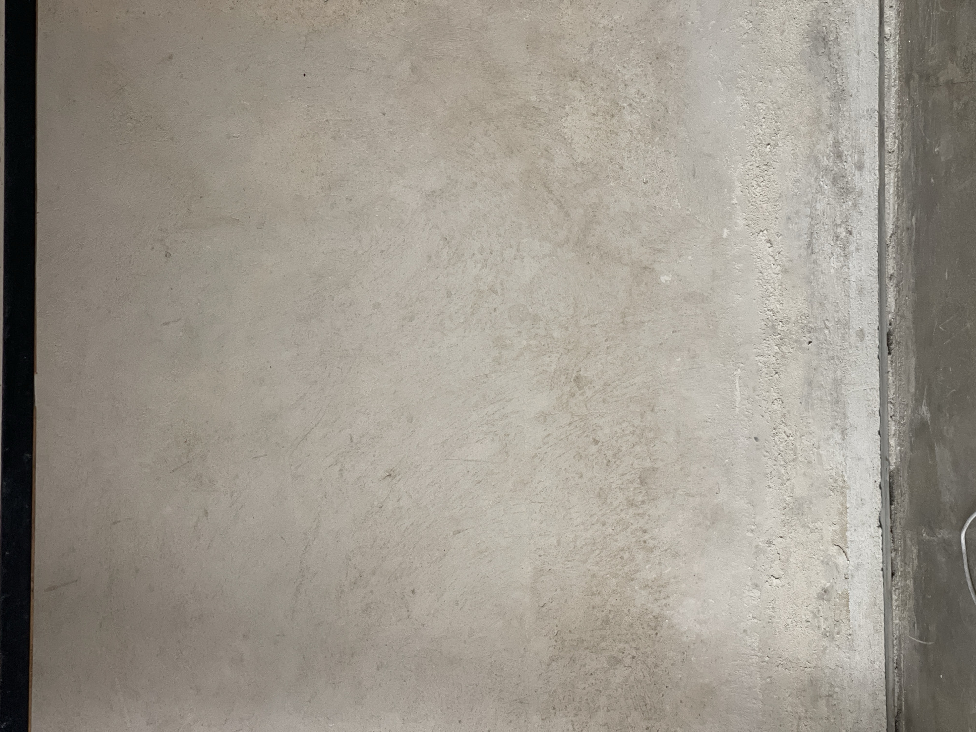 The image shows a section of a concrete floor, likely indoors. The concrete has a textured, slightly uneven surface with subtle variations in color and tone, suggesting possible wear or staining. The left edge of the frame reveals a dark, possibly black, bordering material. The right edge shows a transition to another section of wall or floor with a smoother, grey surface. A small white wire or cable is visible in the lower right corner.
There is no information visible to indicate the location is in North Kuta, Indonesia.