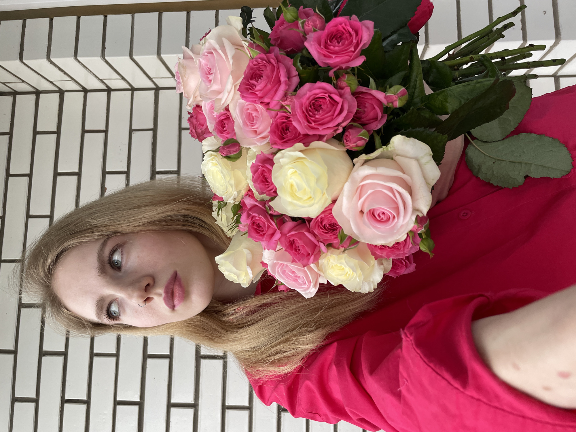 A young woman with light blonde hair and a nose ring is holding a large bouquet of roses in various shades of pink and white. She is wearing a bright pink shirt. The background appears to be a white brick wall with dark grout. The location and city are not discernible in the image.