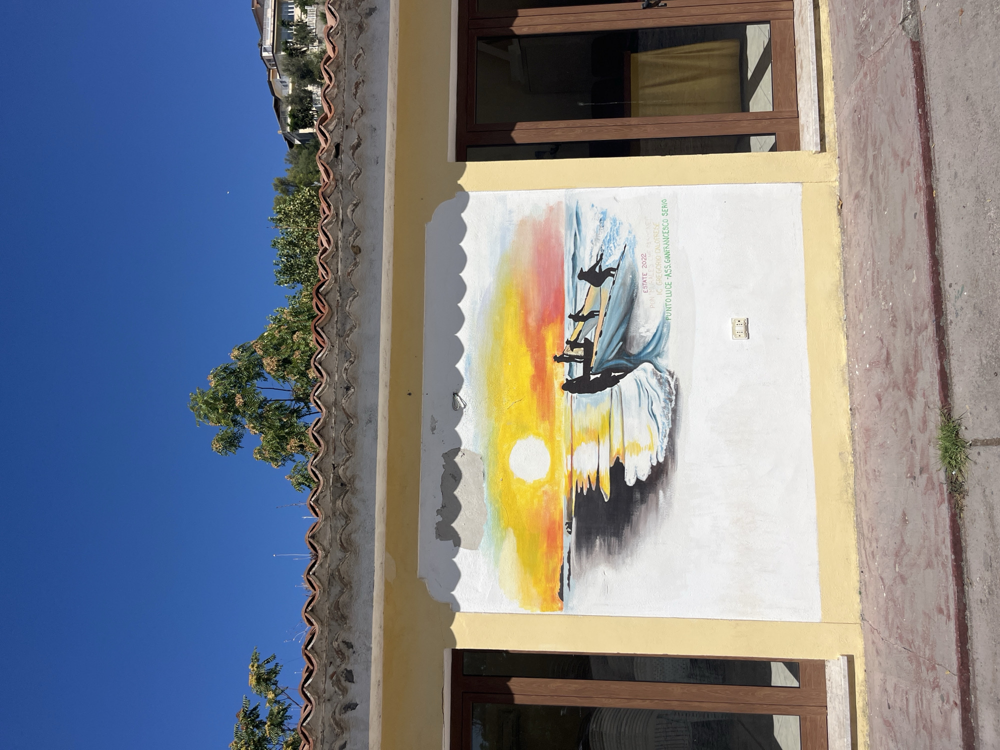 A mural depicting a sunset over the ocean is painted on the exterior of a yellow building with a white trim. The mural shows a group of silhouetted figures in a boat, casting nets into the water. The sky is a vibrant orange and yellow, with the sun prominently displayed. The building has two large glass doors with wooden frames, and a tiled roof. The ground in front of the building is paved with reddish concrete. The scene appears to be in a sunny, outdoor location. The text on the mural reads "ESTATE 2022 PON MURALE DEL PAESAN" and "IC GREGORIO CALOPRESE PUNTO LUCE - ASS. GIANFRANCESCO SERIO". These inscriptions, particularly "PON MURALE DEL PAESAN" which translates to "Bridge Mural of the Villager," and "IC GREGORIO CALOPRESE" which refers to the Gregorio Caloprese Institute, suggest the mural is located in a place associated with the community and a local educational institute. This, combined with the style of architecture and the general setting, is consistent with a location in Scalea, Italy, a coastal town in the Calabria region.