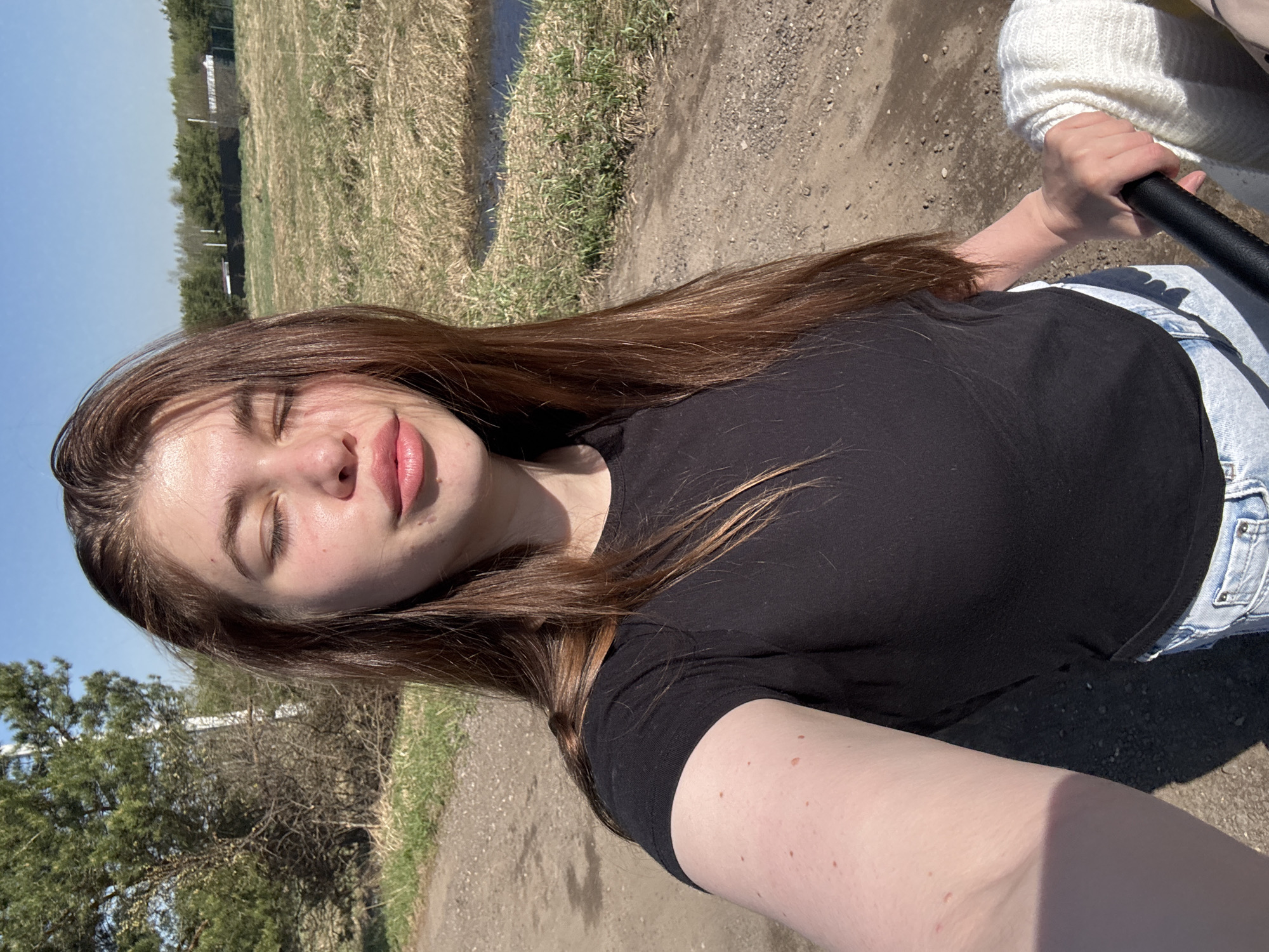 A woman with long brown hair, fair skin, and closed eyes is positioned in the foreground. She wears a black T-shirt and light-wash denim jeans. Her right hand is holding a black handle of a stroller, and a person in a white knit sweater is partially visible in the upper right. The background depicts a rural landscape with a dirt path, dry grass, trees, and a clear blue sky. The location appears to be in the Мещёры region of Russia.