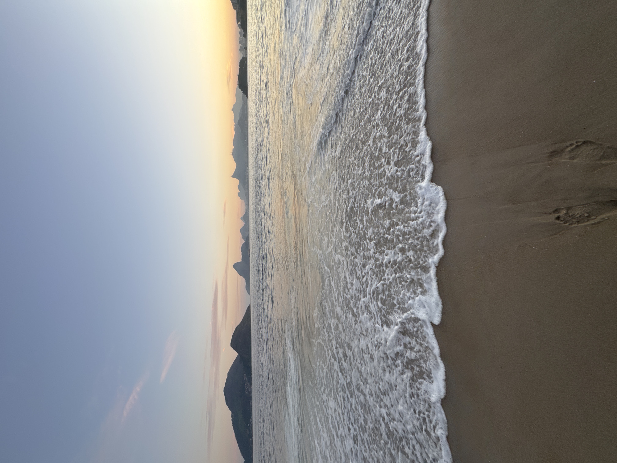 The image captures a serene beach scene in Niterói, Brazil, at sunrise or sunset, given the soft, warm light and pastel hues in the sky. The foreground is dominated by a wet sand beach with a single set of footprints clearly visible, indicating recent human presence. Gentle waves are lapping at the shore, leaving a line of white foam as they recede.
In the middle ground, the calm water of Guanabara Bay stretches out, reflecting the colors of the sky. Beyond the bay, the iconic silhouette of Rio de Janeiro's mountains, including Sugarloaf Mountain and Corcovado (with Christ the Redeemer statue), are visible in the distance. The soft light casts a gentle glow on the landscape, creating a peaceful and picturesque atmosphere. The sky transitions from a pale blue at the top to a warm yellow and orange gradient near the horizon. The overall impression is one of tranquility and natural beauty, characteristic of a dawn or dusk on this Brazilian coastline.