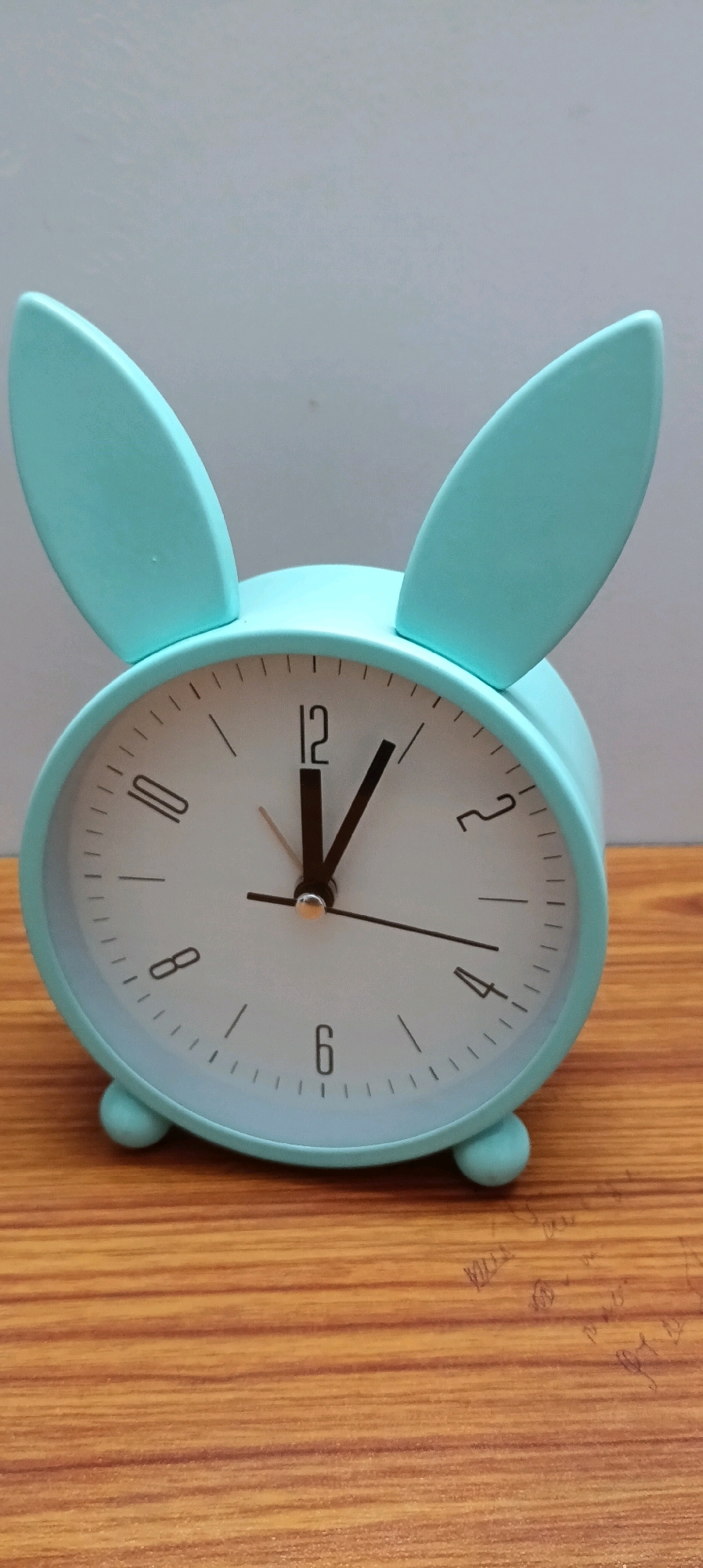 A teal-colored, rabbit-shaped alarm clock with bunny ears is positioned on a wooden surface. The clock face is white with black hour markers and hands. The hour hand points to approximately 11, and the minute hand points to approximately 5. The clock has two small spherical feet. The background is a plain, light gray wall. There are some faint, illegible markings on the wooden surface. The image does not depict any specific location in Udaipur, India, nor does it show any people or actions.