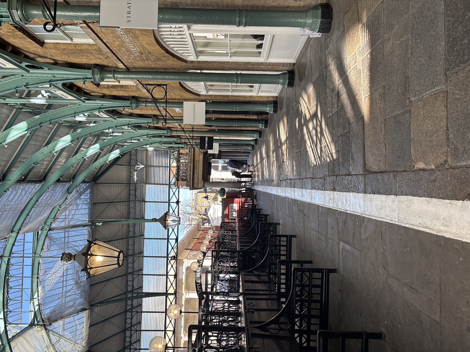 The image depicts an interior view of Covent Garden Market in London, United Kingdom. The market is characterized by a long, covered walkway with a green, arched, glass-paned roof. Architectural details include green support columns with hanging signs for shops such as Strathberry. Rows of black metal seating line the walkway. The floor is stone-paved and features a long white line marking a pathway. Sunlight streams through the roof, creating sharp shadow patterns on the floor. In the distance, a few figures are visible, but details about their actions are unclear. The image appears to be taken during the daytime, with visible sky through the glazed roof.