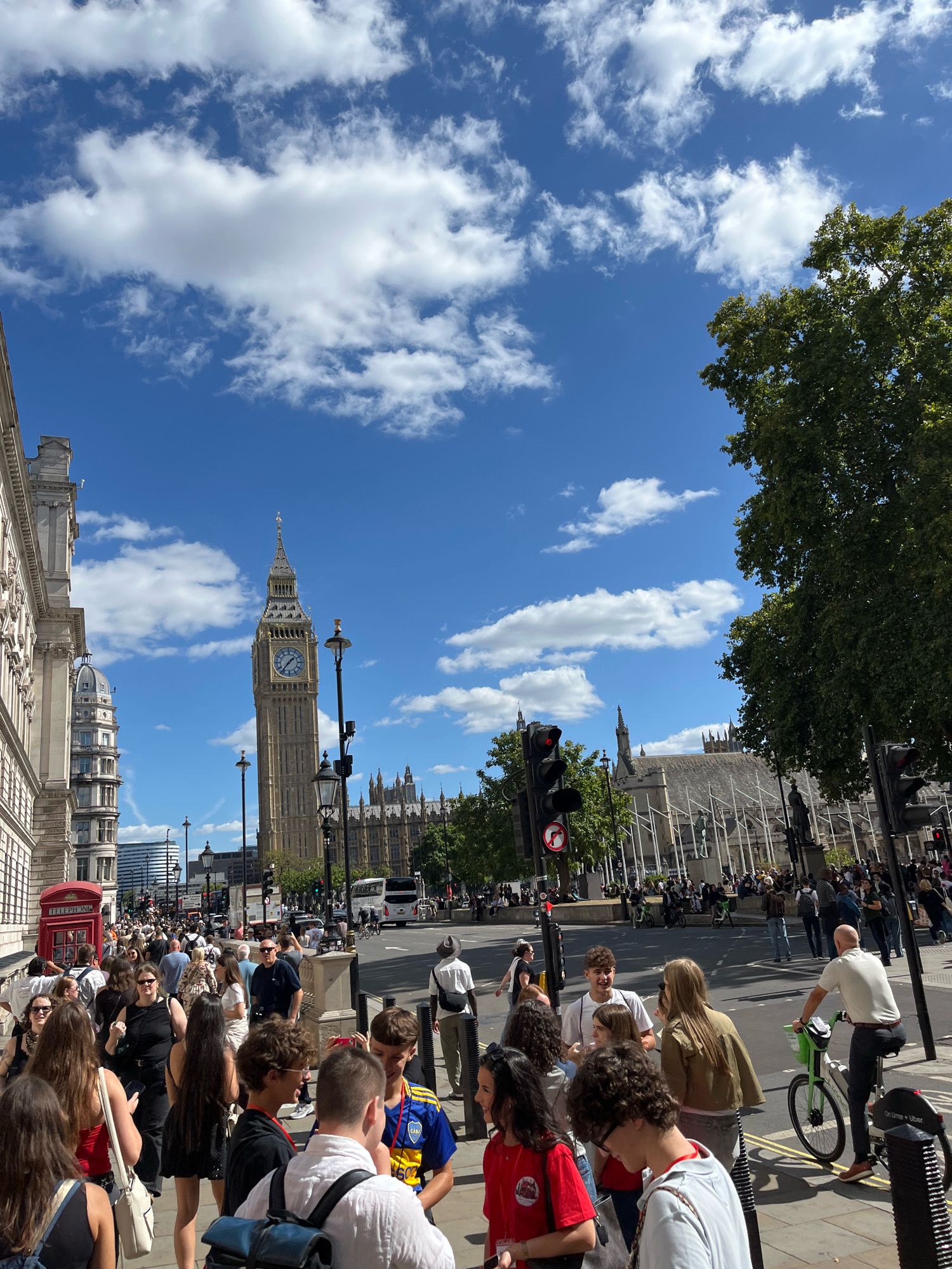 The image captures a bright, sunny day in London, United Kingdom, with the iconic Elizabeth Tower, commonly known as Big Ben, prominently featured. The tower stands tall against a blue sky dotted with fluffy white clouds. In the foreground, a bustling street scene unfolds with a crowd of people walking along the sidewalk and crossing the street.
Several individuals are seen engaging in various activities: some are talking amongst themselves, others are looking at their phones, and a few are taking pictures. A man is cycling on a green bike. On the sidewalk, a classic red British telephone box is visible. Cars, buses, and a bicycle share the road. In the background, the Houses of Parliament and other historic buildings are visible, reinforcing the London setting. The overall atmosphere suggests a lively tourist area on a pleasant day.