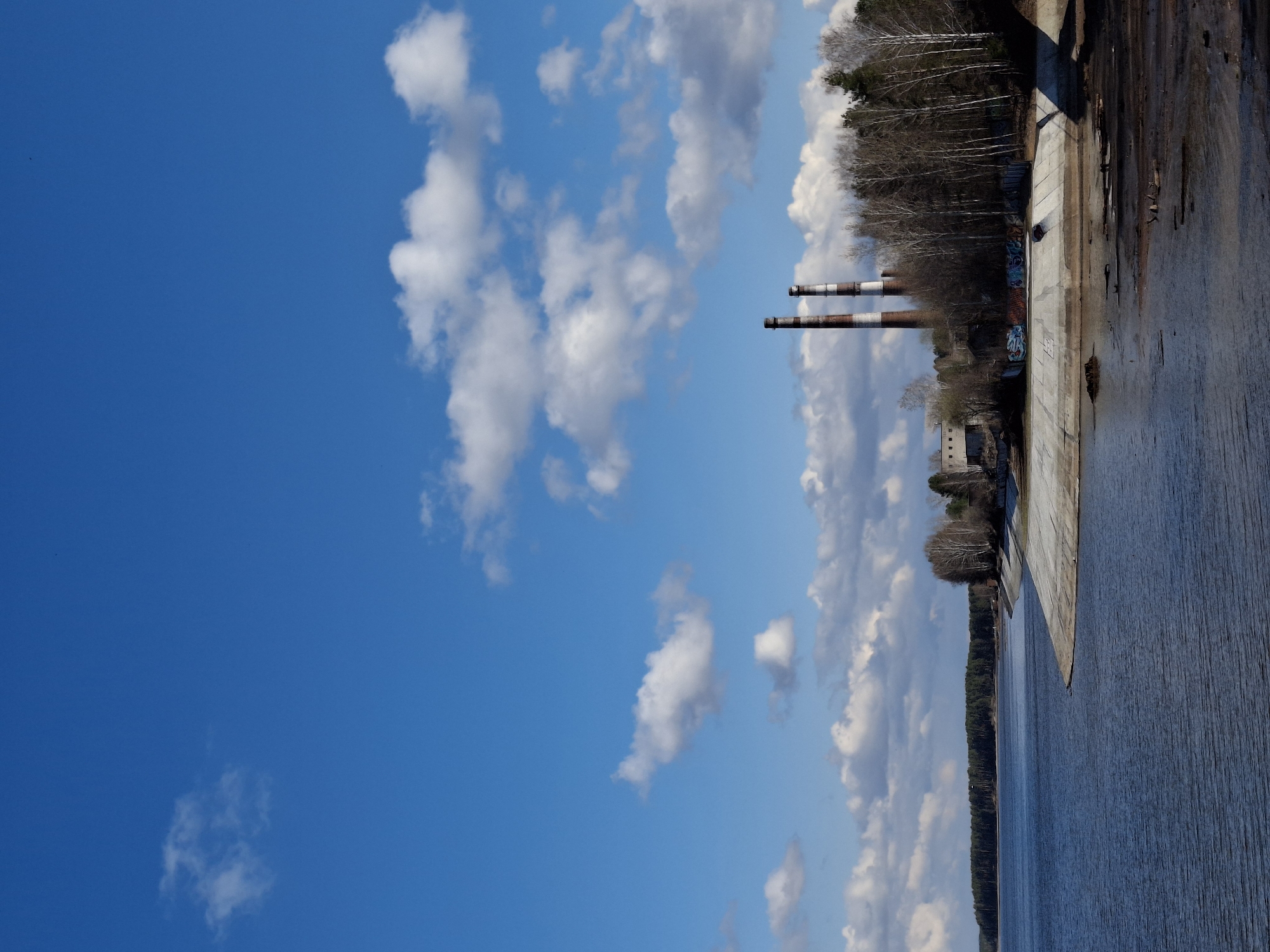 The image shows an area near a body of water, likely a reservoir or lake. A concrete wall or embankment is visible, with the water stretching out in the foreground. On the far side of the water, a line of trees forms the shoreline. Two tall smokestacks rise above the trees, indicating the presence of an industrial facility. Graffiti is visible on the concrete structure near the trees. The sky is blue with scattered white clouds.
Given the prompt's context, this is likely the dam and reservoir area near the city of Chaikovsky, Russia, with the industrial facility potentially being a power plant or manufacturing complex. The image suggests a mixture of natural and industrial elements in the landscape.