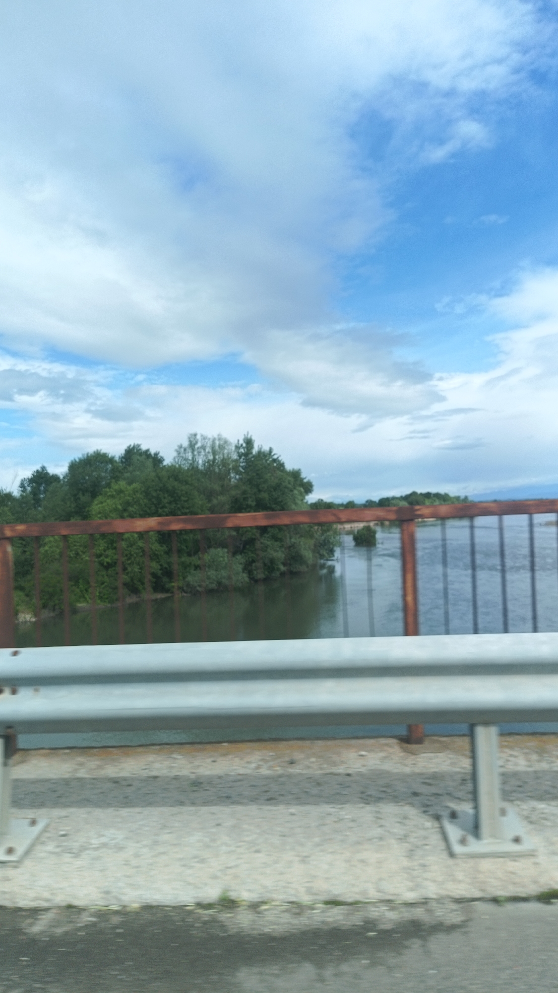 The image shows a view from a bridge over a body of water, likely a river, in Milevo, Bulgaria. The sky is predominantly blue with scattered white clouds. Lush green vegetation covers the riverbank, partially obscured by a rusty, brown metal railing. A grey metal guardrail runs parallel to the road on the bridge, closer to the viewer. The road surface appears wet, possibly from recent rain. No people are visible.