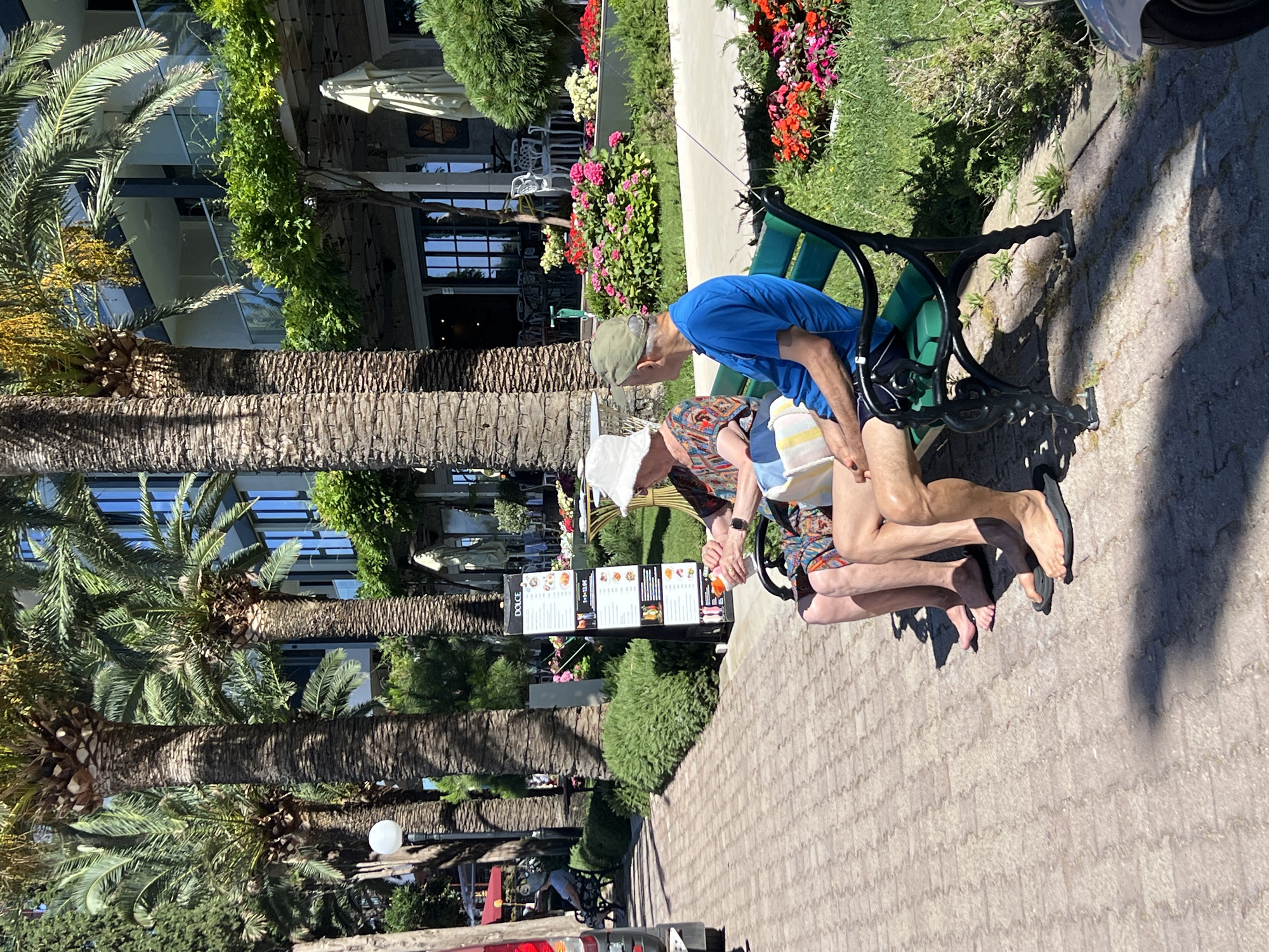 The image depicts an outdoor scene in Rafailovići, Montenegro, with a focus on two elderly individuals seated on a green park bench. The man on the right wears a blue t-shirt and dark shorts, a green cap, and black flip-flops. The woman on the left has a white hat, a patterned dress, and is holding a small orange object. They appear to be sitting close to each other, engaged in conversation or simply resting.
The bench is positioned on a paved walkway constructed with reddish-brown interlocking bricks. Behind the bench, there is a lush green hedge and a vibrant flower bed filled with pink and red flowers. Further in the background, several tall palm trees with rough, textured trunks dominate the view, their fronds casting shadows. Beyond the palm trees, a modern building with multiple levels and reflective windows is visible, suggesting a resort or hotel setting. There are also tables and chairs from an outdoor dining area. The overall atmosphere suggests a warm, sunny day in a coastal tourist area.