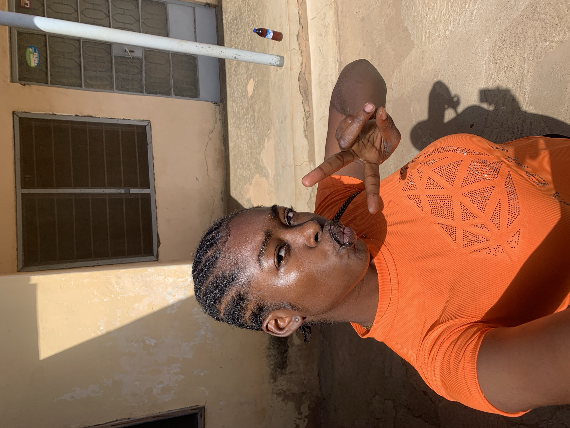 A person is posing outdoors, likely in Rafin Par, Nigeria, given the image's origin. The individual, wearing an orange top with geometric patterns, is displaying a hand gesture. A small bottle floats in the background near a building with barred windows. The ground is a mix of concrete and dirt, partially in shadow. The shadow of a hand holding a camera is visible.