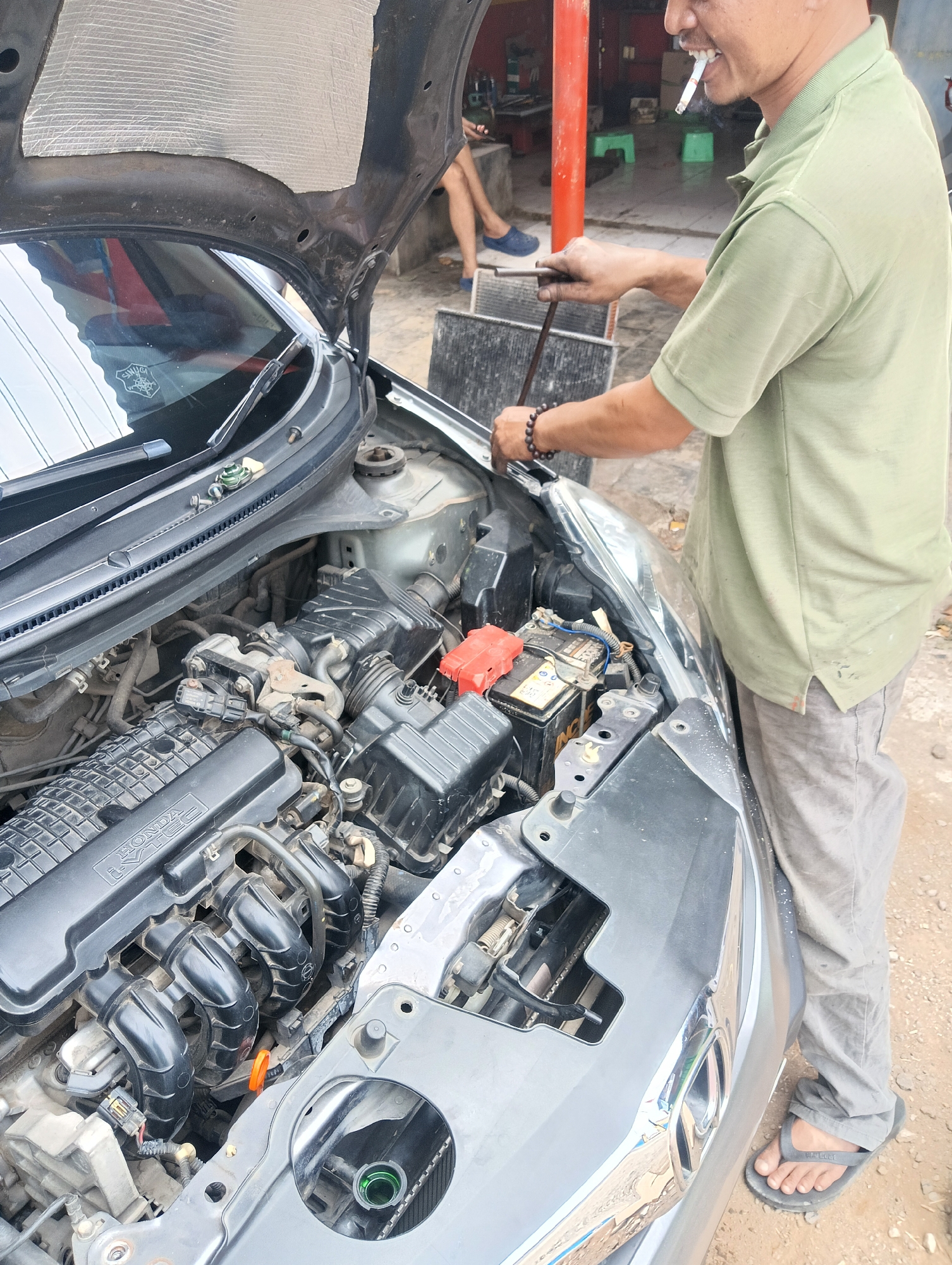 In Manggari, Indonesia, a mechanic with a cigarette in his mouth is working on the engine of a car with the hood open. He is wearing a light green polo shirt and grey pants. The car's engine bay is visible, showing various components. In the background, another person is seated, and a red pillar is present. The ground is dirt, and there are some tools scattered around.
