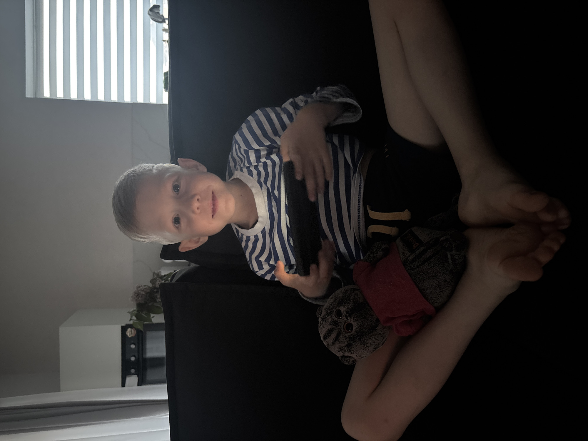 A young child, possibly a toddler, is seated in a darkened room. The child is holding a black electronic device. The environment is dimly lit. A stuffed animal rests near the child's legs. A window with blinds is visible in the background. The child is wearing a striped shirt. A microwave oven and bouquet of flowers are visible. The location is Chaikovsky, Russia.
