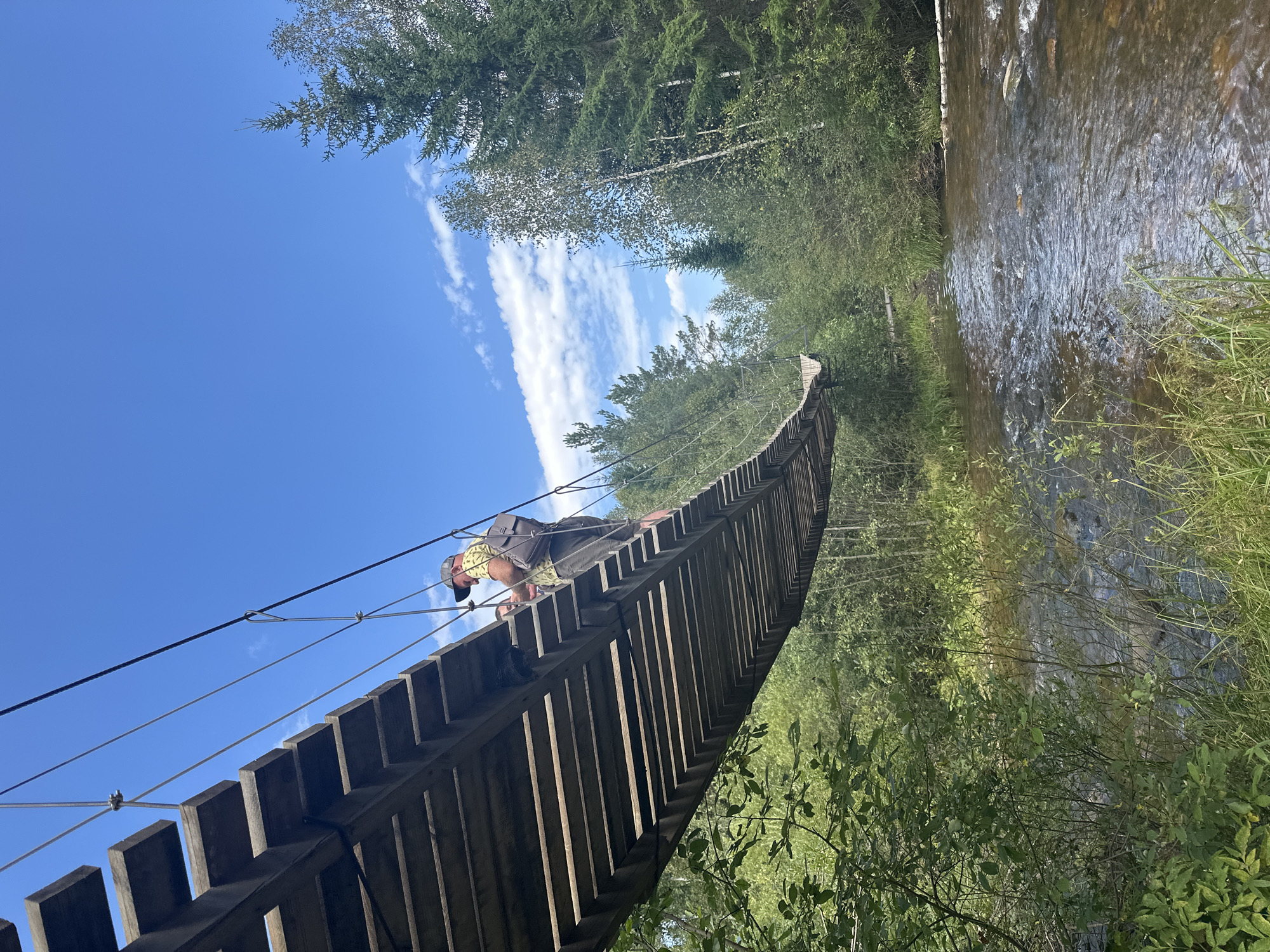 An overhead low-angle shot captures a person on a wooden suspension bridge crossing over a shallow, clear river. The bridge is constructed with narrow wooden planks and supported by cables. The person, wearing a hat, a patterned shirt, and dark pants, is walking away from the camera. Lush green trees and foliage border the riverbanks, with taller coniferous trees in the background. The sky above is a bright blue with scattered white clouds.