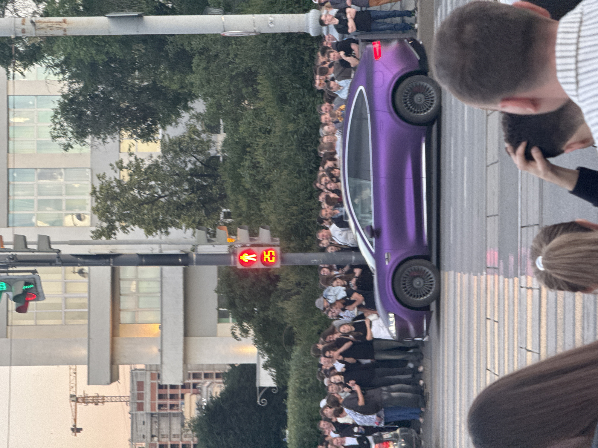 The image depicts a street scene in Yekaterinburg, Russia, likely near the Yekaterinburg City Administration building given the architectural style. A crowd of people is gathered, appearing to be waiting at a pedestrian crossing. A traffic light displays a red walking figure and the number "25," indicating a countdown for pedestrians. A striking metallic purple car is prominently featured in the foreground, stopped at the crossing. In the background, a construction crane is visible, suggesting ongoing development in the area. The overall atmosphere appears to be one of anticipation or observation of the car or an event.