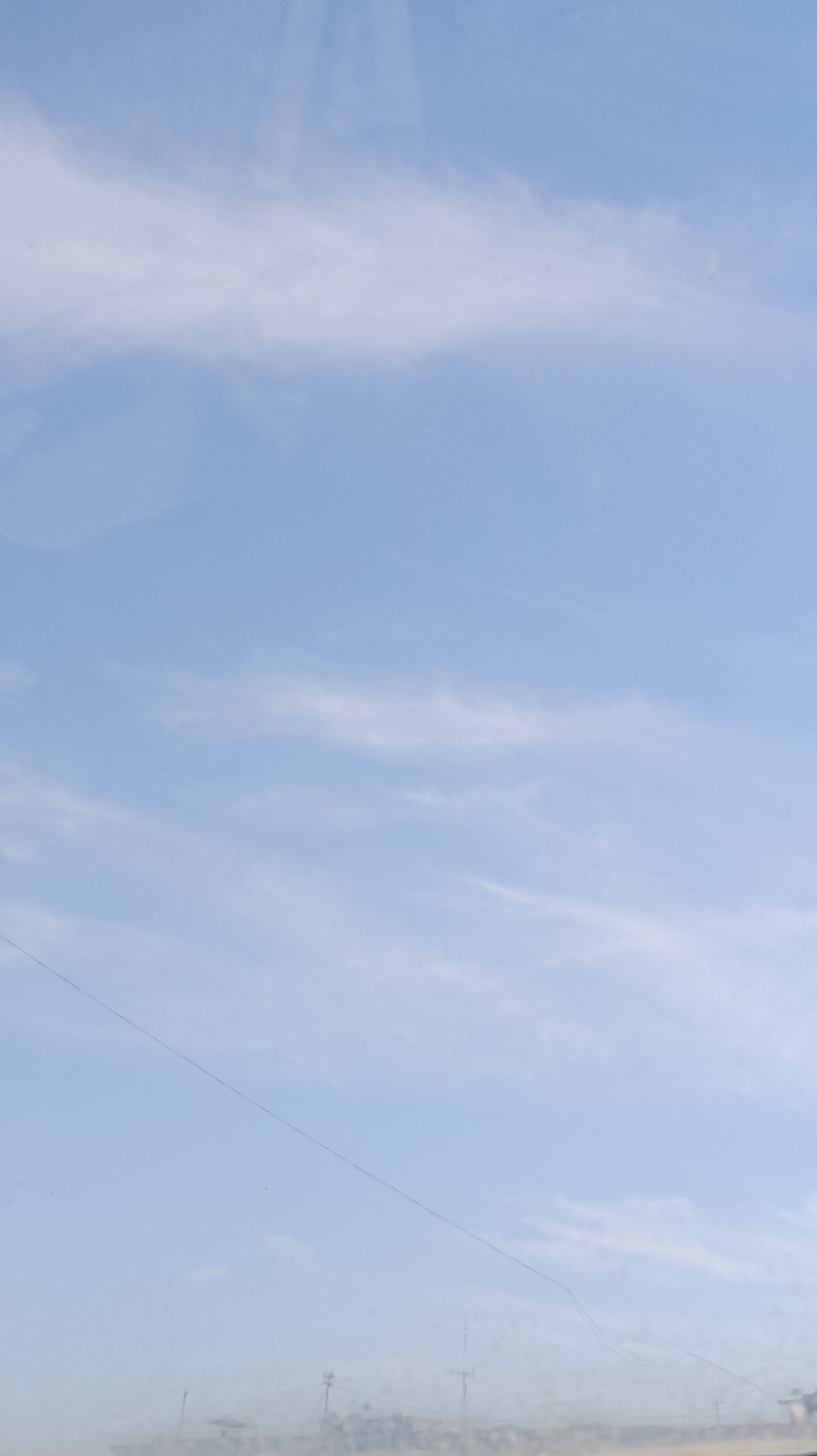The image shows a pale blue sky with wispy white clouds. In the lower portion of the image, a faint, hazy line of what appears to be a city skyline is visible, with a few indistinct structures that could be antennas or poles. A thin, dark line, possibly a wire or a crack in a surface, runs diagonally across the sky from the middle left towards the lower right. The overall impression is of an overcast or hazy day with a slightly muted color palette.