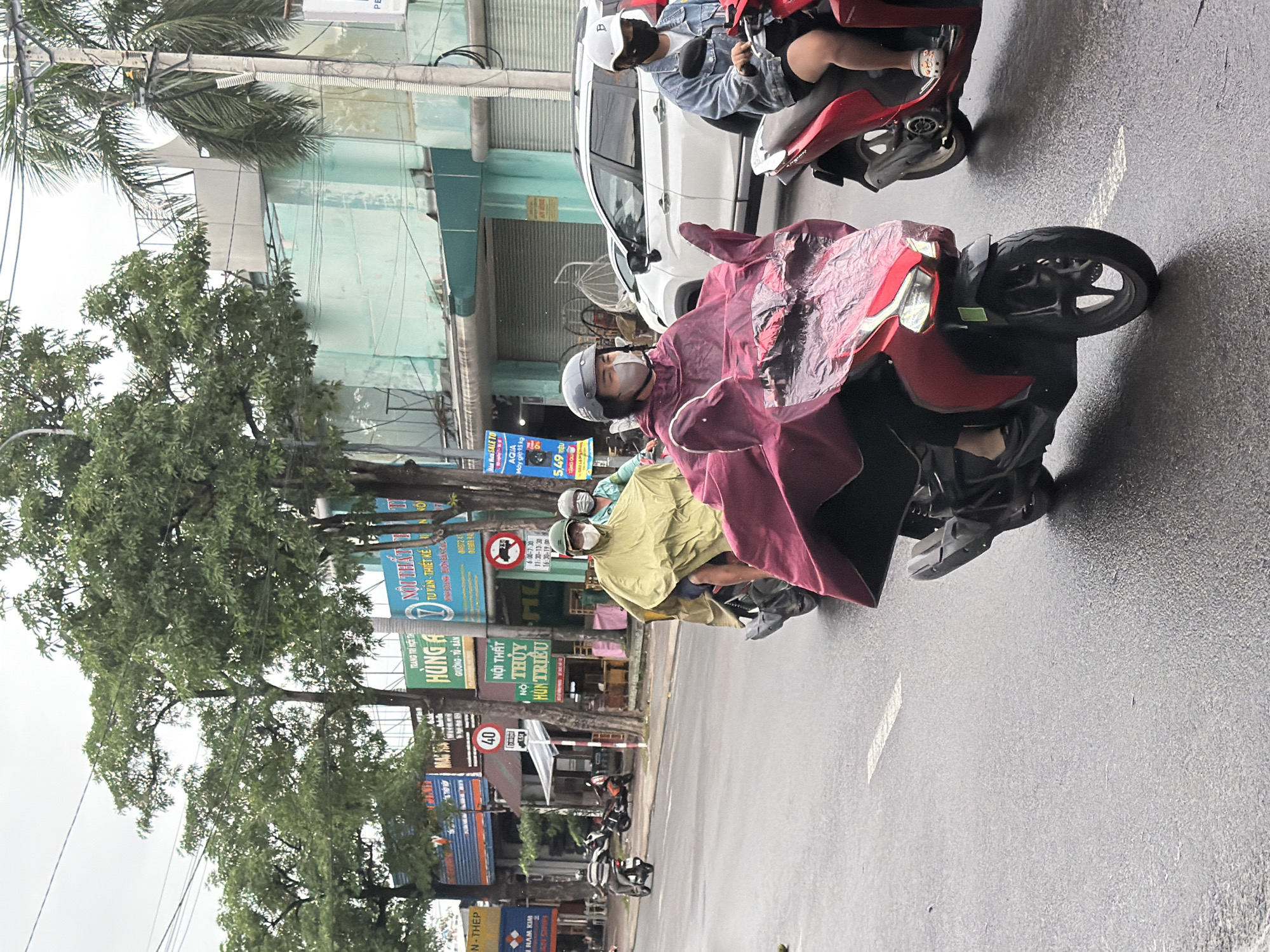 The image depicts a street scene in Nha Trang, Vietnam, characterized by wet asphalt and an overcast sky, suggesting recent or ongoing rain. Several individuals are seen riding motorcycles, many of whom are wearing protective rain gear, including ponchos and helmets.
In the foreground, a person wearing a red poncho is riding a red motorcycle. They are wearing a grey helmet and a face mask. The poncho covers a significant portion of the motorcycle and the rider. Behind them, another motorcycle with two occupants is visible, with the rider wearing a light yellow poncho and the passenger wearing a blue jacket and a white helmet.
Further in the background, other motorcycles and a white car can be seen on the road. Buildings with various signage line the street, some of which appear to be shops or businesses. Trees with lush green foliage are also present along the roadside, contributing to the urban landscape. The overall atmosphere is one of daily life continuing despite the wet weather conditions.