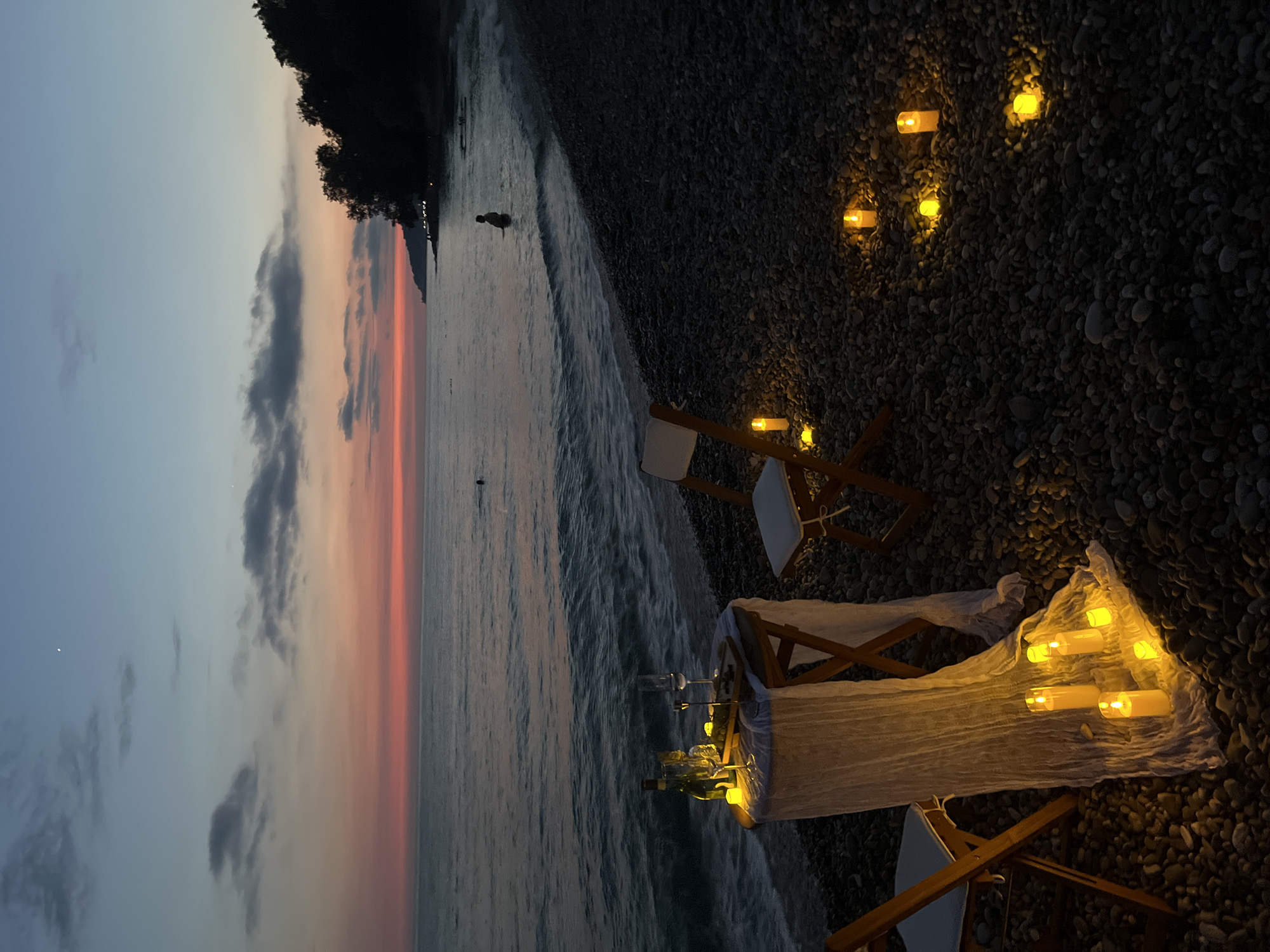 This image depicts a romantic dinner setup on a pebble beach in Sochi, Russia, at dusk. A small, round wooden table is covered with a white, sheer cloth and set with two wine glasses, a bottle of wine, and a small platter of food, possibly cheese and grapes. Several battery-operated candles illuminate the table and the surrounding beach. Two folding chairs, with white cushions, are placed at the table. The sea is calm, with gentle waves lapping at the shore. In the background, two figures are visible in the water, and a treeline with distant lights borders the coast. The sky displays a gradient of colors, from a pale blue at the top to shades of pink and orange near the horizon, indicating the sunset. The overall atmosphere is serene and intimate, suggesting a private romantic experience.