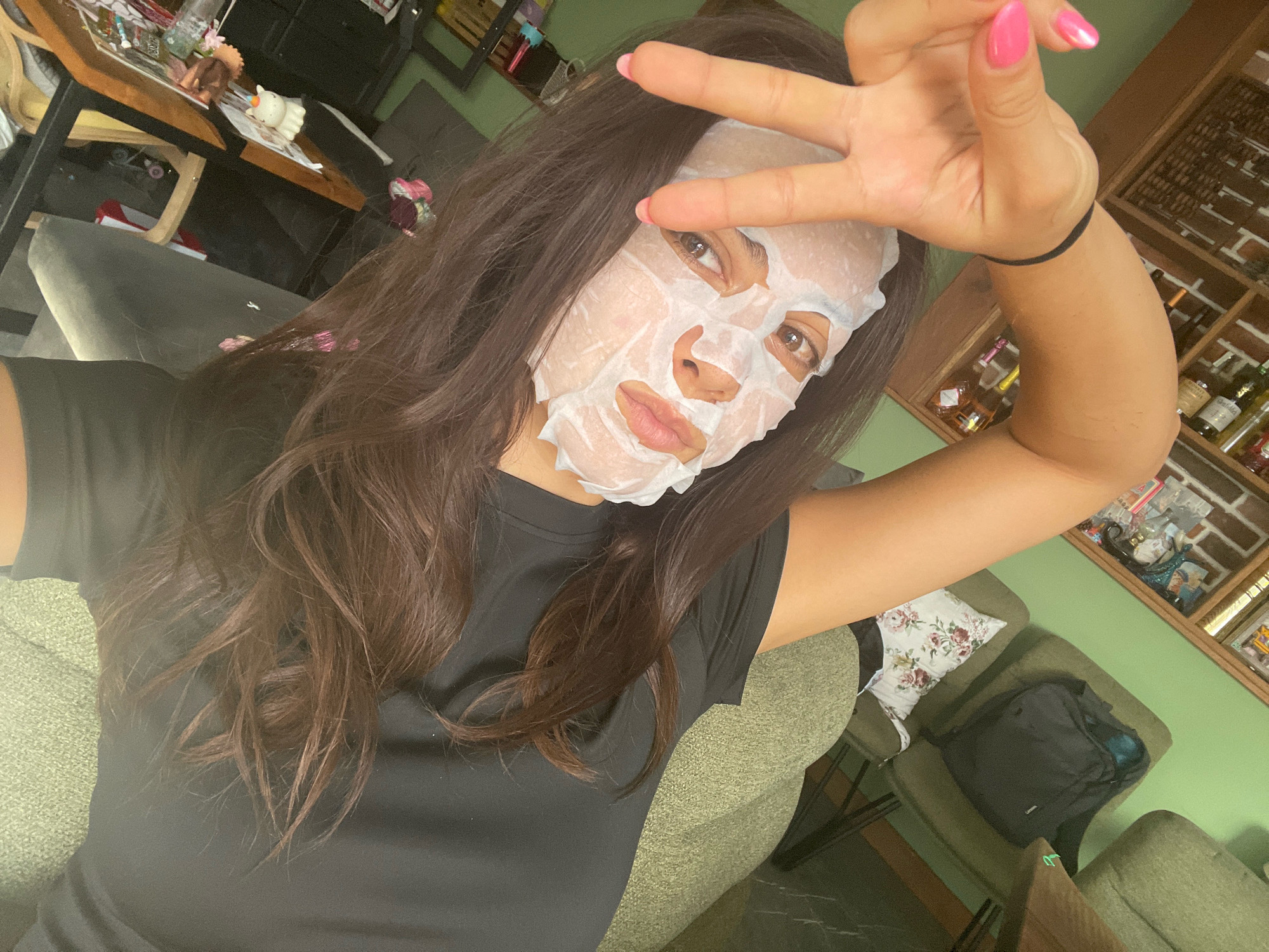 A woman with long, dark brown hair is taking a selfie while wearing a white sheet mask on her face. She is making a peace sign with her right hand, which has bright pink nail polish. Her left arm is raised and bent, with a black hair tie around her wrist. She is wearing a black t-shirt. The background shows a room with green walls, a wooden shelf unit filled with bottles and decorative items, and a textured green sofa. A table with various items on it is visible to the left. The scene appears to be indoors.