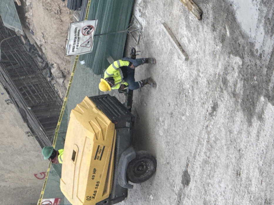 Two construction workers are present in the scene. One, wearing a yellow hard hat and safety vest, appears to be working on a piece of yellow machinery. The other worker wears a green hard hat and appears to be inspecting the machine. A sign is present on the ground, as well as concrete forms and steel rebar structures. The ground appears to be made of cement.