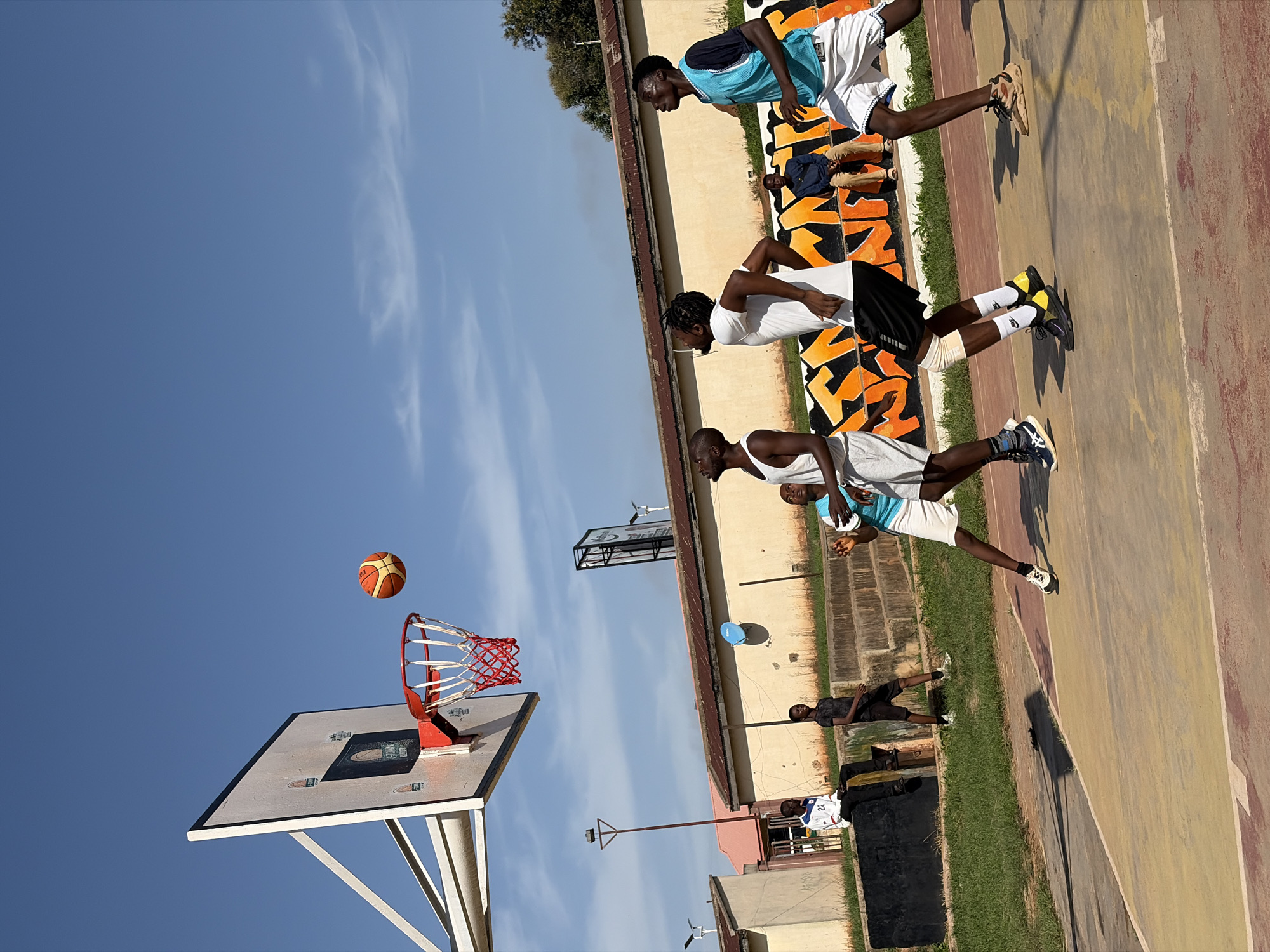 In Jos, Nigeria, on a sunny day, five young men are playing basketball on an outdoor court. A basketball is in mid-air, heading towards the hoop. Four players are actively engaged in the game. One player in a white tank top and grey shorts is jumping, while another in a white t-shirt and black shorts is looking up, possibly preparing to rebound. A third player, wearing a blue tank top and white shorts, is also looking up. The fourth active player is out of frame. Two other young men are observing the game; one is sitting on a concrete ledge in the foreground, wearing a white shirt with the number 22. Another is standing in the background, wearing a dark shirt and shorts. The court is made of a worn, multi-colored surface, with a concrete ledge and grass surrounding it. Behind the court, there is a building with an orange and black graffiti mural. The sky is clear blue with a few wispy clouds.