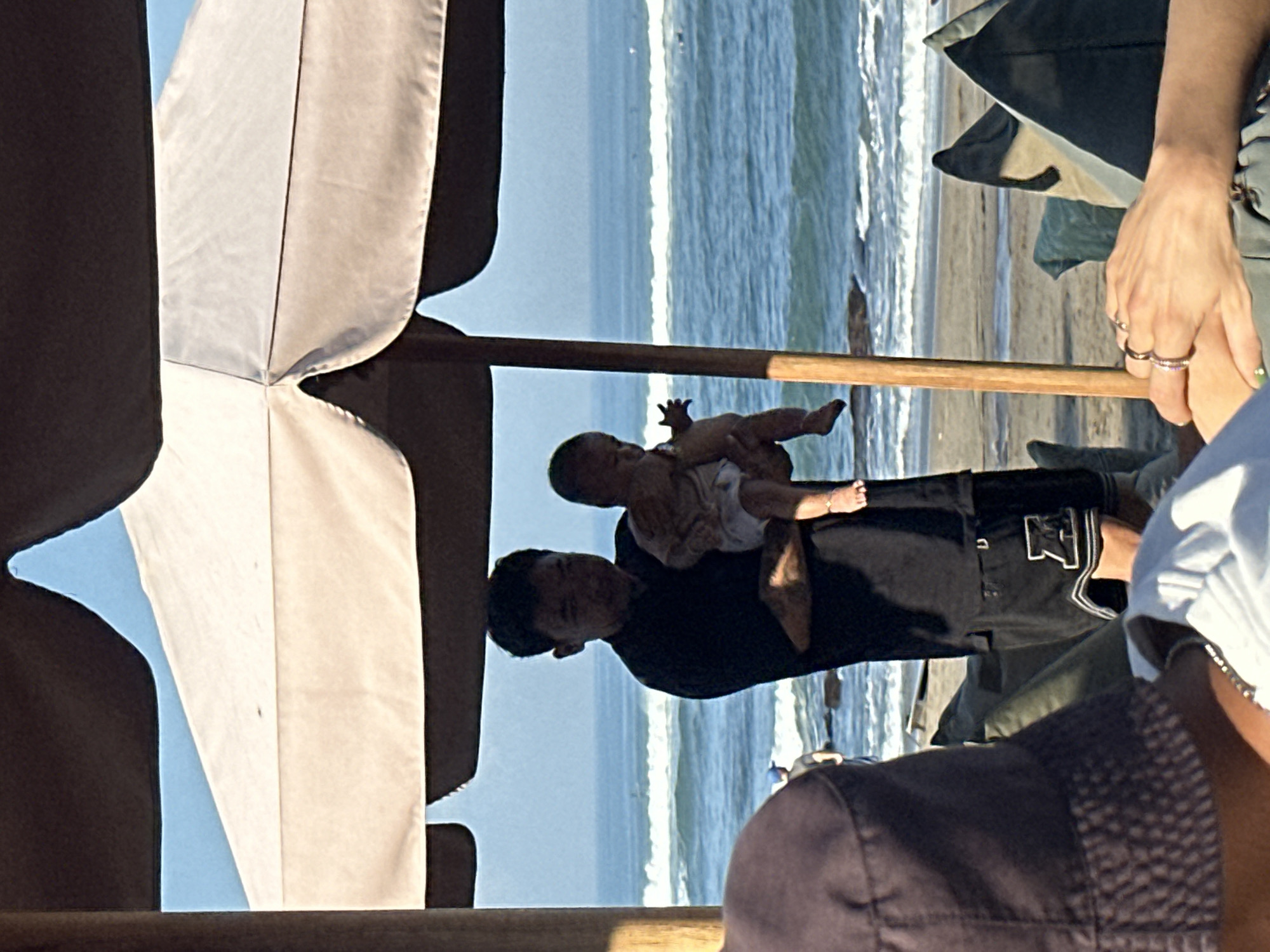 A man is holding a baby on a beach in North Kuta, Indonesia. The man is wearing a black t-shirt and black shorts with a white logo. The baby is wearing a white diaper. They are standing under a large umbrella, with the ocean in the background. In the foreground, a person wearing a dark hat and a person wearing a white shirt are visible. The beach appears to be sandy, with some rocks in the water. The sky is blue and clear.