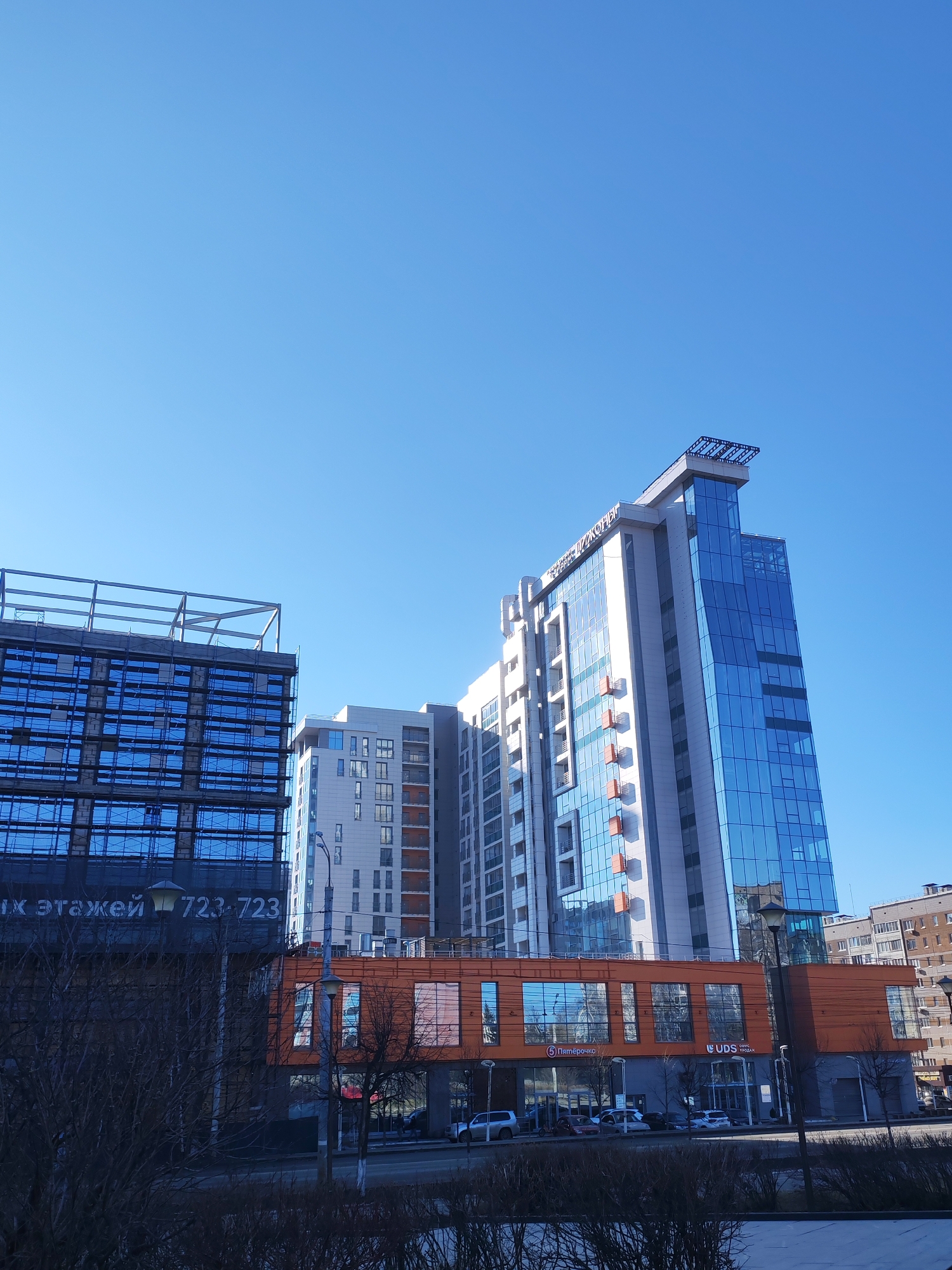 The image captures a low-angle view of modern architecture in Izhevsk, Russia, against a clear blue sky. Three prominent buildings dominate the frame. On the left is a building under construction, covered with blue reflective sheeting and metal scaffolding. Adjacent to it is a lighter-colored residential building with balconies. The tallest structure is a multi-story building featuring a glass facade with an attached orange building at street level. The orange building houses businesses, and street-level signage indicates Russian writing. A street with cars is visible in front of the buildings, partially obscured by barren trees and bushes in the foreground. The buildings appear to be near the center of the city.