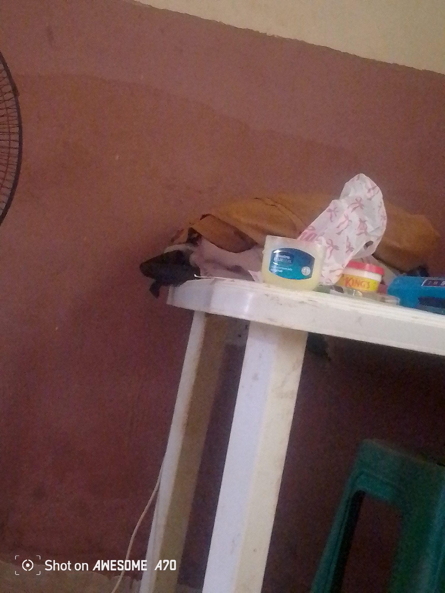 The image shows a close-up view of a white plastic table with various items on it. On the table, there is a blue and white tub of Vaseline, a red and yellow tub with the word "KINGS" visible, and a blue electronic device. Behind the table, there is a brown wall and what appears to be fabric or clothing. To the left of the image, the edge of a black fan is visible. The scene is indoors, with the lighting suggesting it could be daytime, though the lack of windows or natural light sources makes it difficult to determine definitively. The overall impression is of a simple, domestic setting, likely in Jalingo, Nigeria, as indicated by the text watermark at the bottom left which reads "Shot on AWESOME A70". No people or specific activity are depicted, but the presence of personal items suggests a lived-in space. The environment appears somewhat cluttered but functional.