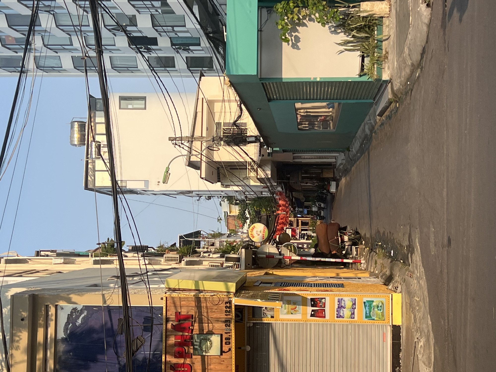 The image captures a narrow street scene in Nha Trang, Vietnam. The street is flanked by buildings of varying heights and architectural styles. High-rise apartments dominate the left side, with numerous windows visible. Electrical wires crisscross the sky, adding to the urban clutter. On the right, a mix of lower buildings includes shops with roller shutters and buildings with green awnings. The road is asphalt, showing some signs of wear and tear along the edges. Debris is scattered on the street and along the curbside. In the distance, activity with tables, chairs and awnings indicate a restaurant or food stall area. The overall impression is of a busy, somewhat chaotic urban environment.