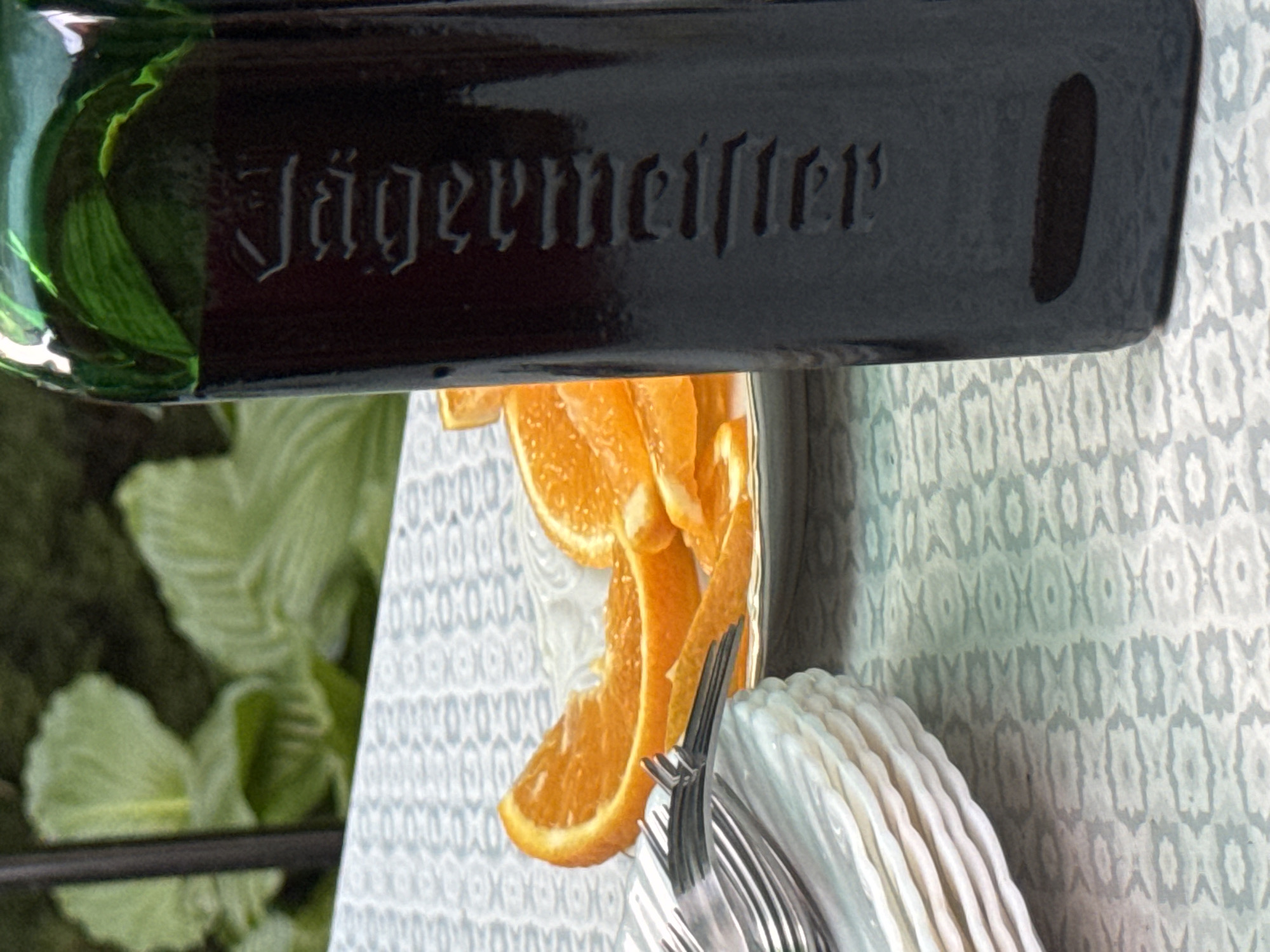 The image shows a close-up of a Jägermeister bottle standing on a patterned tablecloth. The bottle is dark green glass with the brand name "Jägermeister" embossed vertically on its side. In the background, out of focus, are slices of orange on a plate, a stack of white plates, and some greenery. The scene appears to be outdoors, possibly in a garden setting. There is no information or visual evidence in the image to suggest any connection to the city of Pribrezhnyi, Russia.