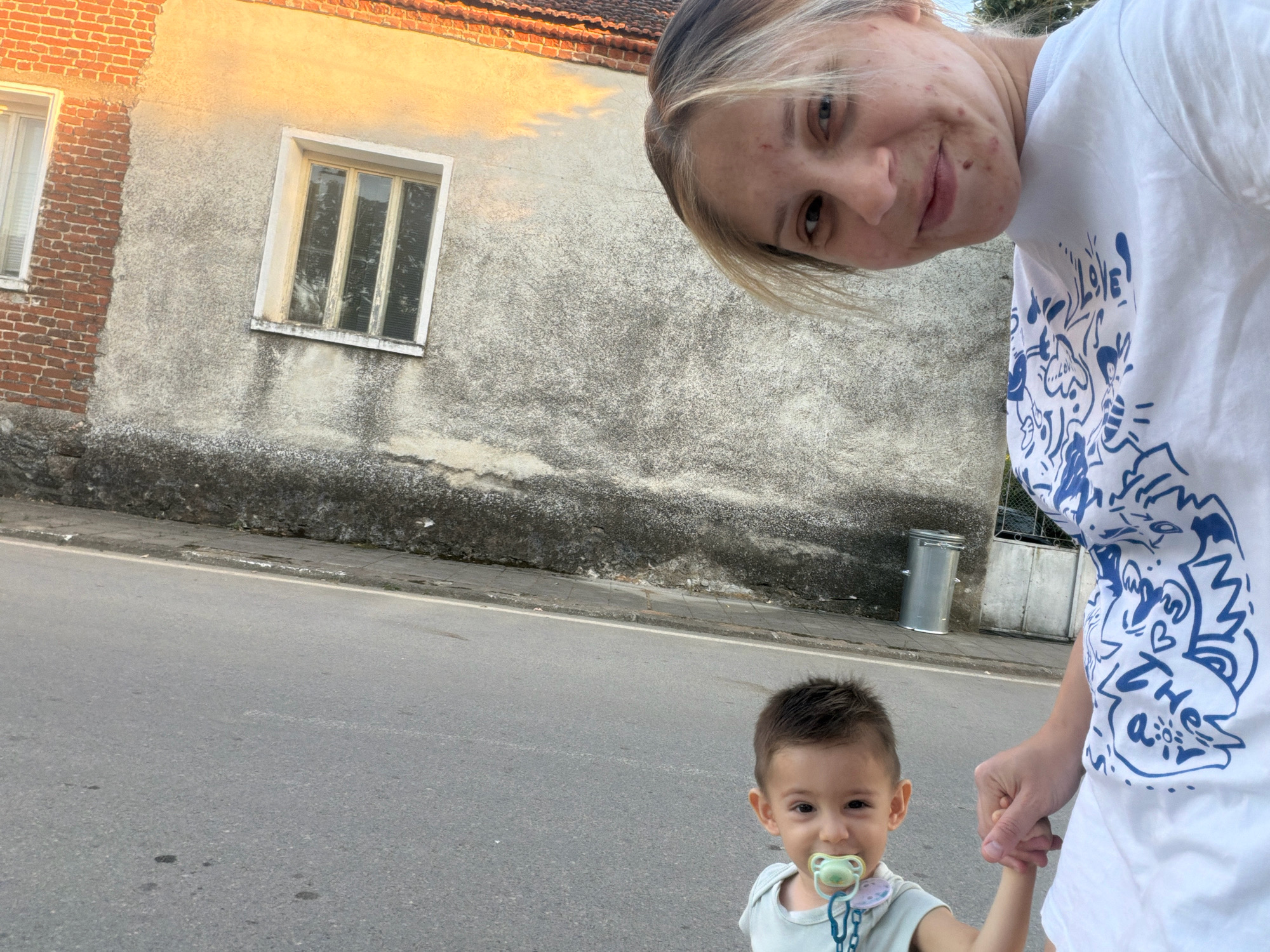In an outdoor urban setting, a young child with a pacifier is being held by the hand by an adult. The child, with dark hair and brown eyes, wears a light-colored shirt. The adult, partially visible, has blonde hair and is wearing a white t-shirt with blue graphics. They are standing on a paved road. In the background, a building with a textured wall and a brick section is visible, along with a window and a trash can. The lighting suggests it is either morning or evening.
