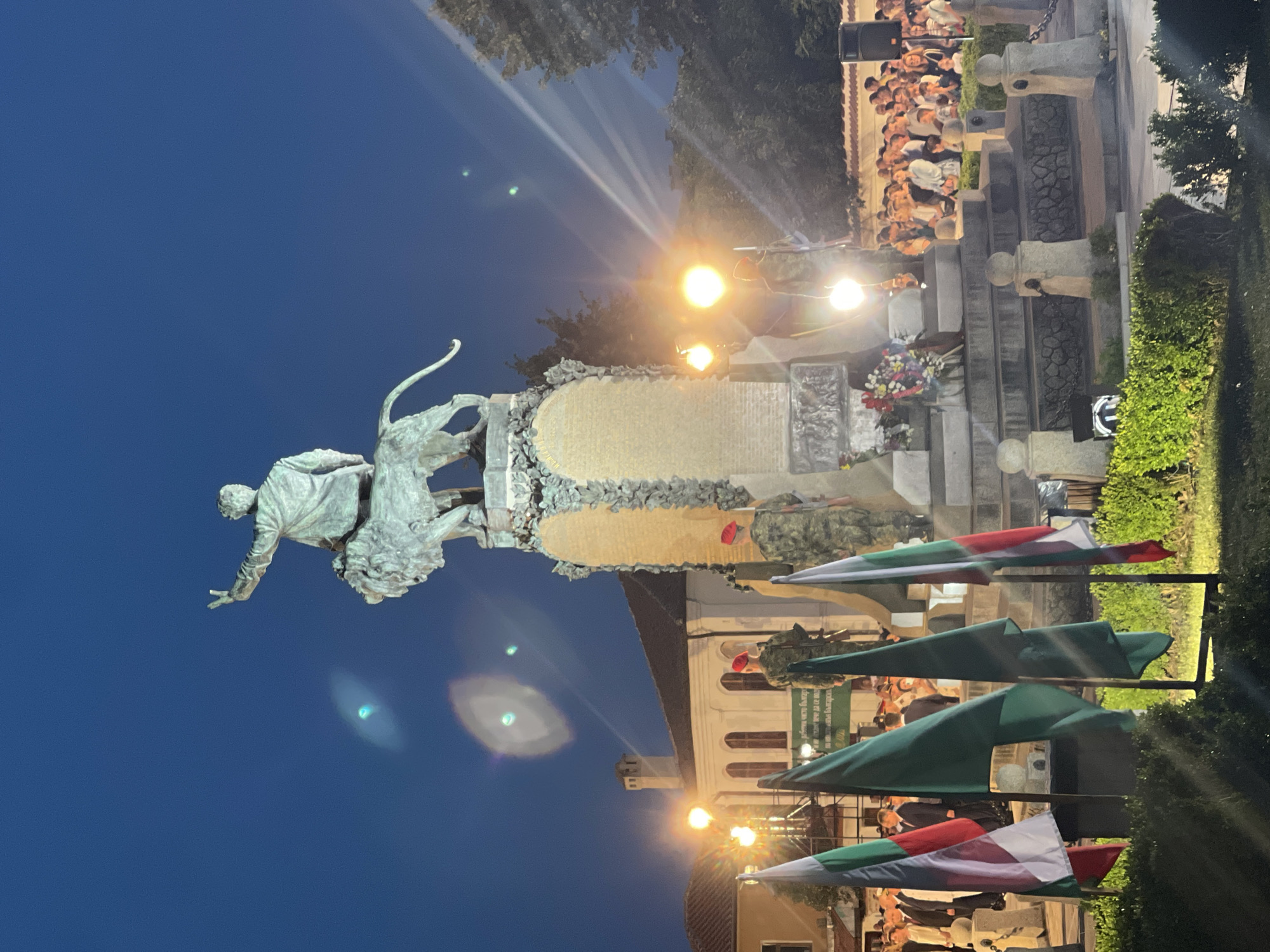 The image depicts an outdoor evening ceremony in front of a monument in Karlovo, Bulgaria. The monument features a statue of a figure on a lion, with intricate carvings and a pedestal. Several Bulgarian flags are prominently displayed in the foreground, with green foliage surrounding the base of the monument. A crowd of people is gathered, observing the ceremony. Two individuals in military uniforms with red hats stand at attention near the monument. Several lights illuminate the scene, creating beams of light in the dark sky. The sky is a deep blue, indicating it is dusk or nighttime. The architecture of a building with arched windows is visible behind the monument. The overall scene suggests a commemorative event or celebration.