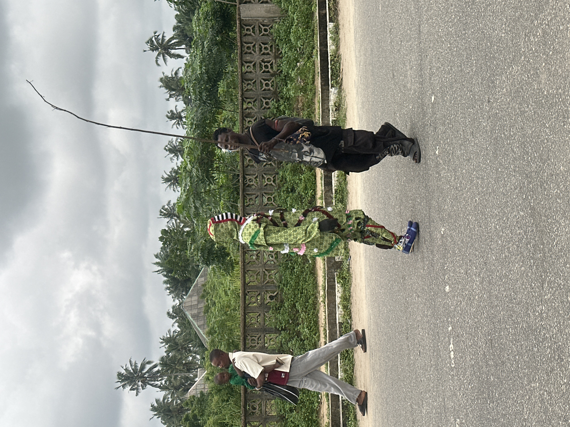 Three individuals are walking on a paved road in Badagry, Nigeria. On the left, a man wearing a cream-colored shirt, maroon undershirt, and grey pants carries a small child dressed in green. In the center, a person in a green costume adorned with pink and white embellishments walks towards the right. On the right, a man in a black t-shirt and dark cargo pants carries a long stick over his right shoulder. He has a bag slung across his chest. Behind the road, a decorative concrete fence borders an area with lush green vegetation and palm trees. In the background, two pyramid-shaped structures with grey roofs are visible among the trees. The sky is overcast with grey clouds.