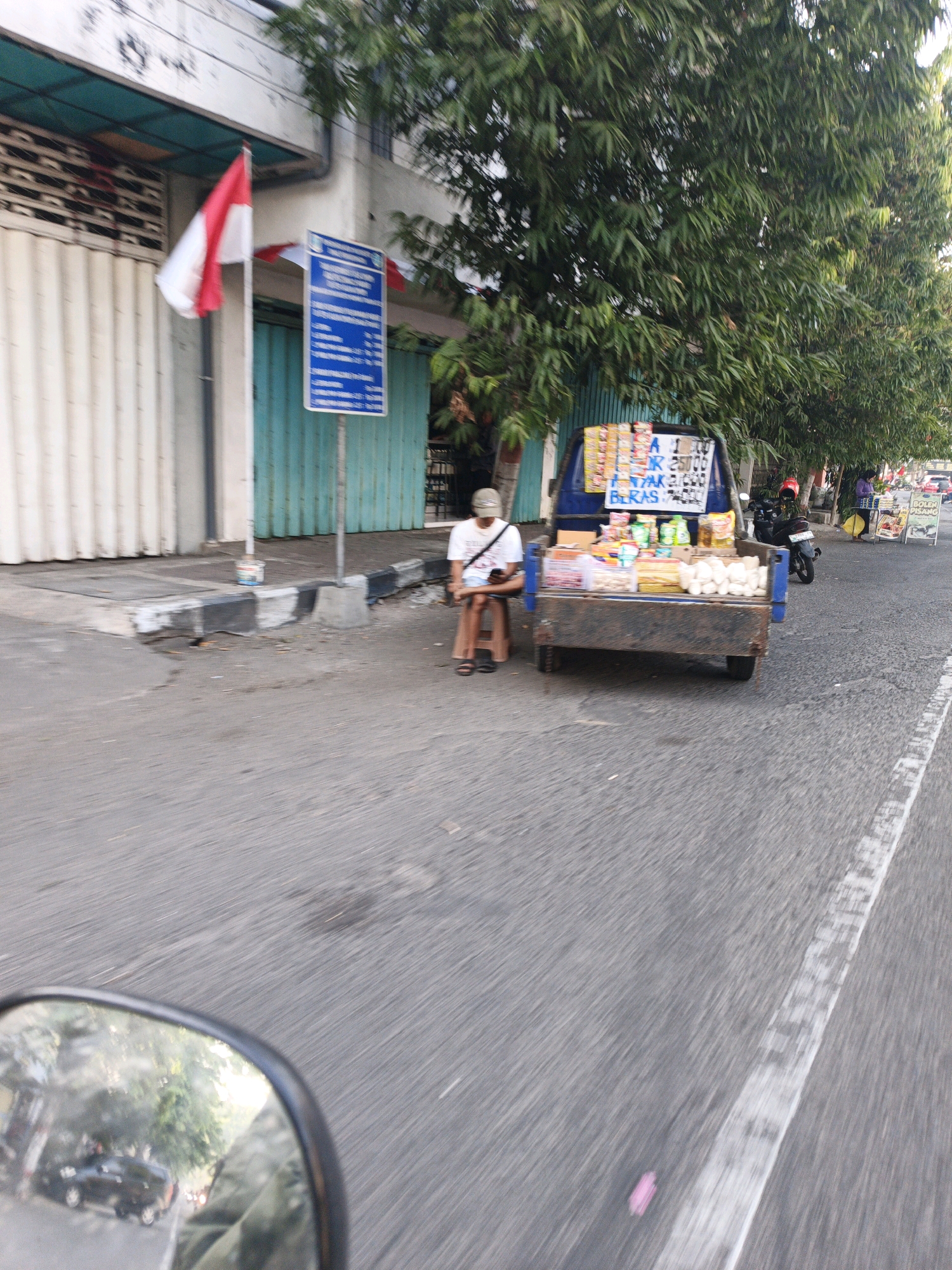 The image shows a street scene in Mojokerto, Indonesia, with a mobile food vendor's cart. The vendor, a person wearing a white t-shirt and a cap, sits on a small stool beside the cart, which is loaded with various packaged snacks and food items. The cart appears to be a modified pickup truck. In the background, a building with a white and teal facade is visible, along with a red and white Indonesian flag displayed on a pole. A blue sign with white text is also present near the building. Trees line the street, providing shade. A motorcycle with a red helmet is parked behind the food cart. Further down the street, other vendors and shops can be seen, along with passing vehicles. The scene is captured from a moving perspective, likely from a vehicle, as indicated by the motion blur and the reflection in the mirror in the foreground which shows a street with trees and a car.