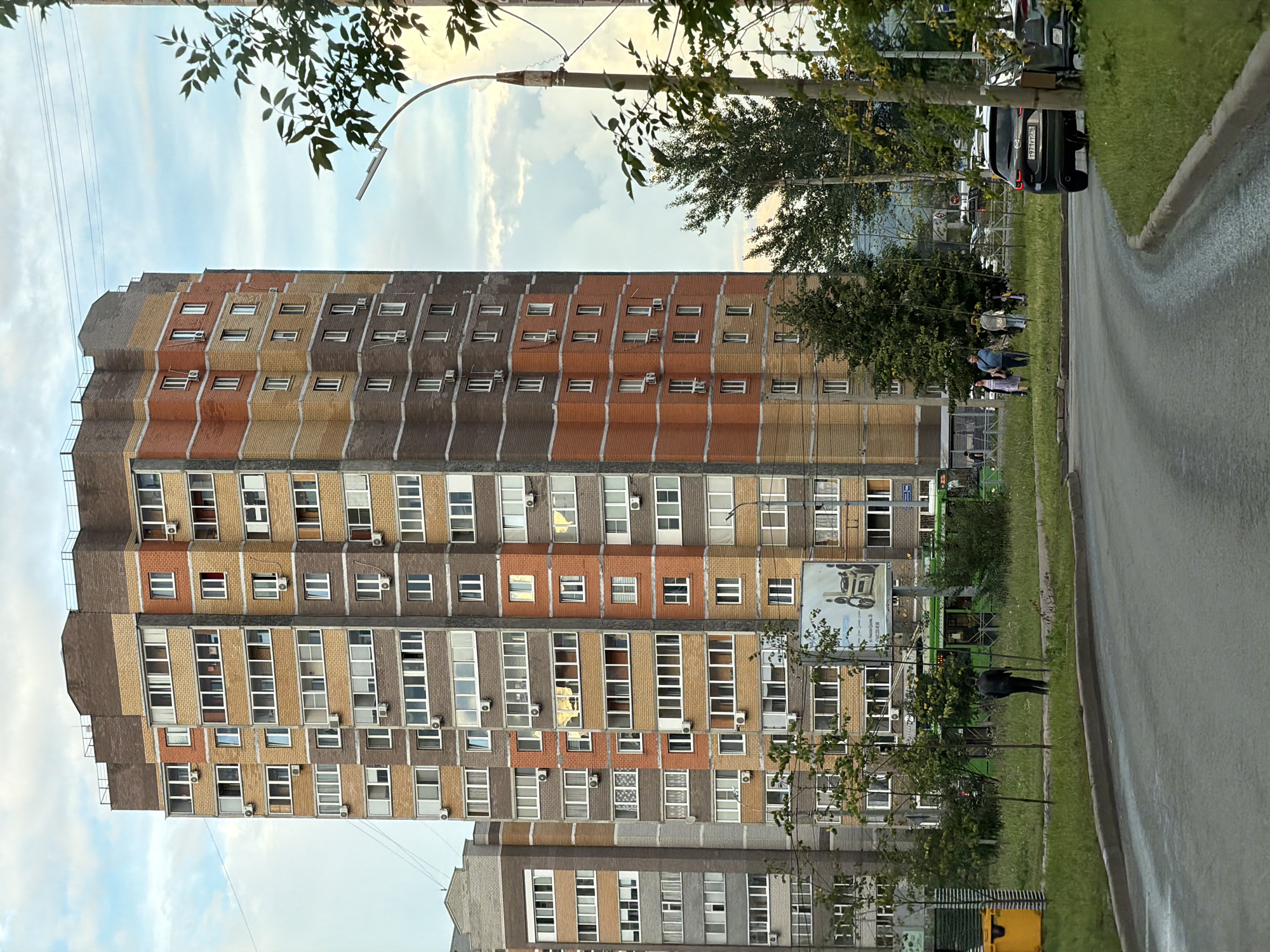 The image shows a tall, multi-story apartment building constructed with brick in shades of brown, orange, and grey. The building features numerous windows and air conditioning units. In the foreground, a paved road curves to the right, with a grassy median and sidewalk. A dark-colored SUV is parked on the right side of the road, partially obscured by foliage. Several people are walking on the sidewalk near the building, and a green tram or bus is visible in the background. Trees and a streetlamp are also present. A billboard with an advertisement is attached to the side of the apartment building. The sky is cloudy. The scene appears to be a residential area within the city of Kazan, Russia.