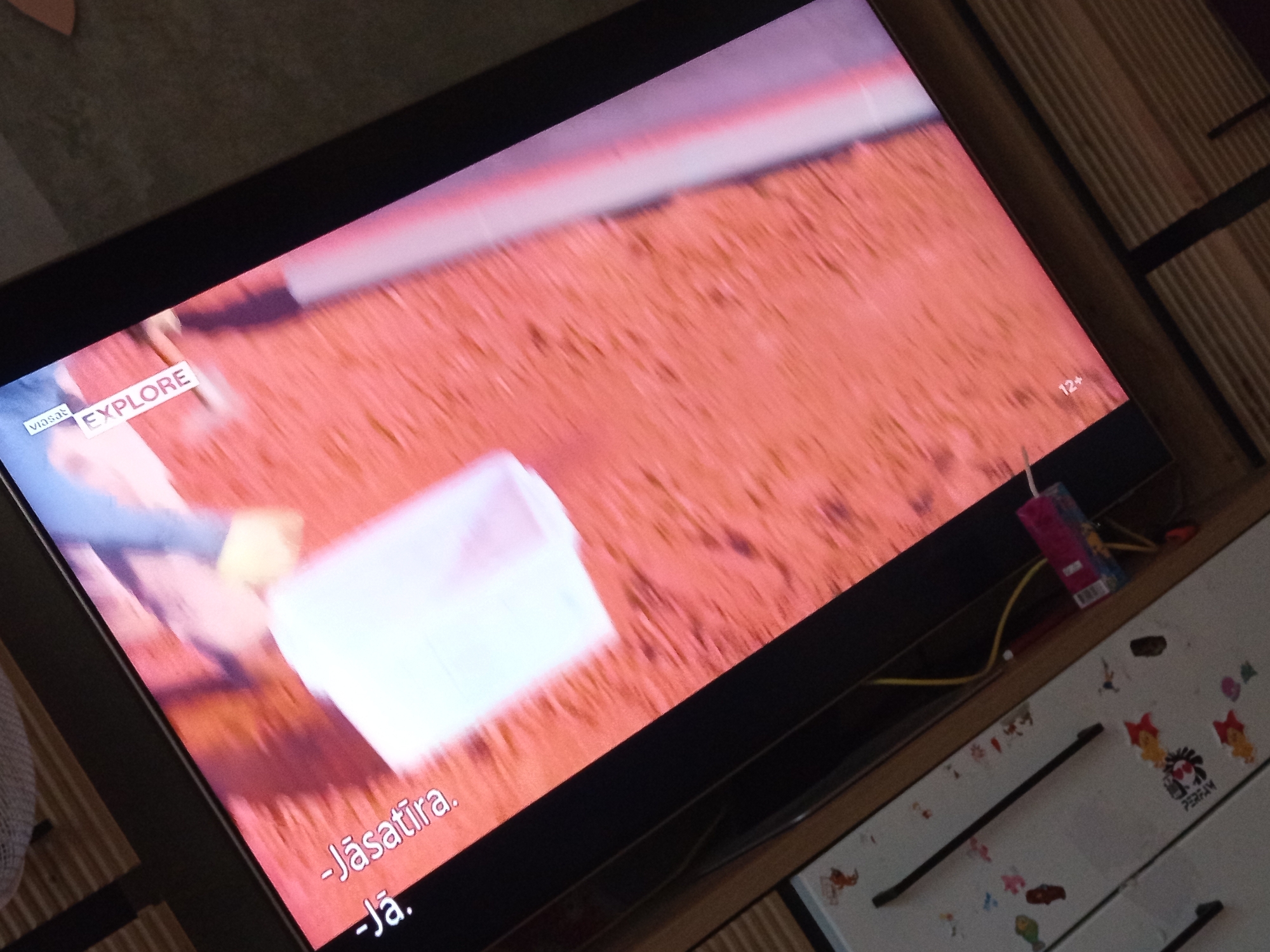 The image shows a television screen displaying a scene with text overlays. On the screen, a pinkish-red textured surface is visible, possibly representing ground or a fabric. A person, mostly out of focus, is seen on the left side of the screen, wearing blue clothing and interacting with a white translucent object. Towards the bottom of the screen, Latvian subtitles are displayed, reading "-Jāsātira." and "-Jā.". In the upper left corner of the screen, the word "EXPLORE" is visible, preceded by what appears to be a logo for "Viasat". On the right side of the screen, a "12+" age rating is displayed. The television is placed on a wooden surface, which has some stickers and what appear to be drawer handles. The overall lighting suggests an indoor setting. There is no information in the image to suggest a location in Zīlāni, Latvia.