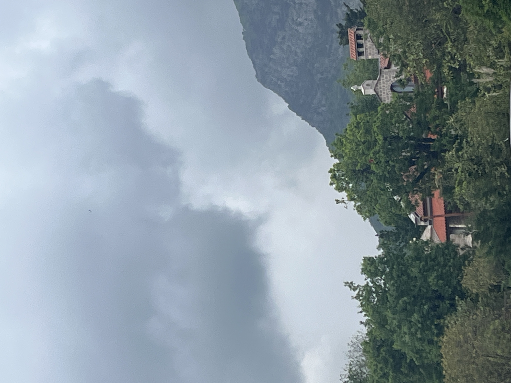 This image captures a serene, mountainous landscape in Budva, Montenegro, under an overcast sky. The scene is dominated by a large, rugged mountain in the background on the right, its dark, textured slopes partially veiled by atmospheric haze or distant clouds.
The sky, filling the left and central portions of the frame, is covered with dense, light to dark grey clouds, indicating a cloudy or potentially rainy day. This contributes to a muted and diffused lighting across the entire scene.
In the midground and foreground, lush green trees with full foliage are abundant, suggesting a vibrant natural environment. Nestled within these trees, on the lower slopes or adjacent hills, are several buildings with distinct architectural features. One prominent structure appears to be a bell tower or campanile, possibly part of a church or monastery, featuring multiple arched levels, stone-like construction, and what looks like a cross at its apex. Further below and to the right, another building with a reddish-brown tiled roof is visible, also partially obscured by the surrounding greenery. The architecture, with its tiled roofs and stone elements, is typical of the Mediterranean and Balkan regions.
No people or specific activities are visible, emphasizing the tranquility and natural beauty of the location. The time of day cannot be precisely determined due to the heavy cloud cover, but the overall atmosphere is calm and peaceful. There is no visible text in the image.