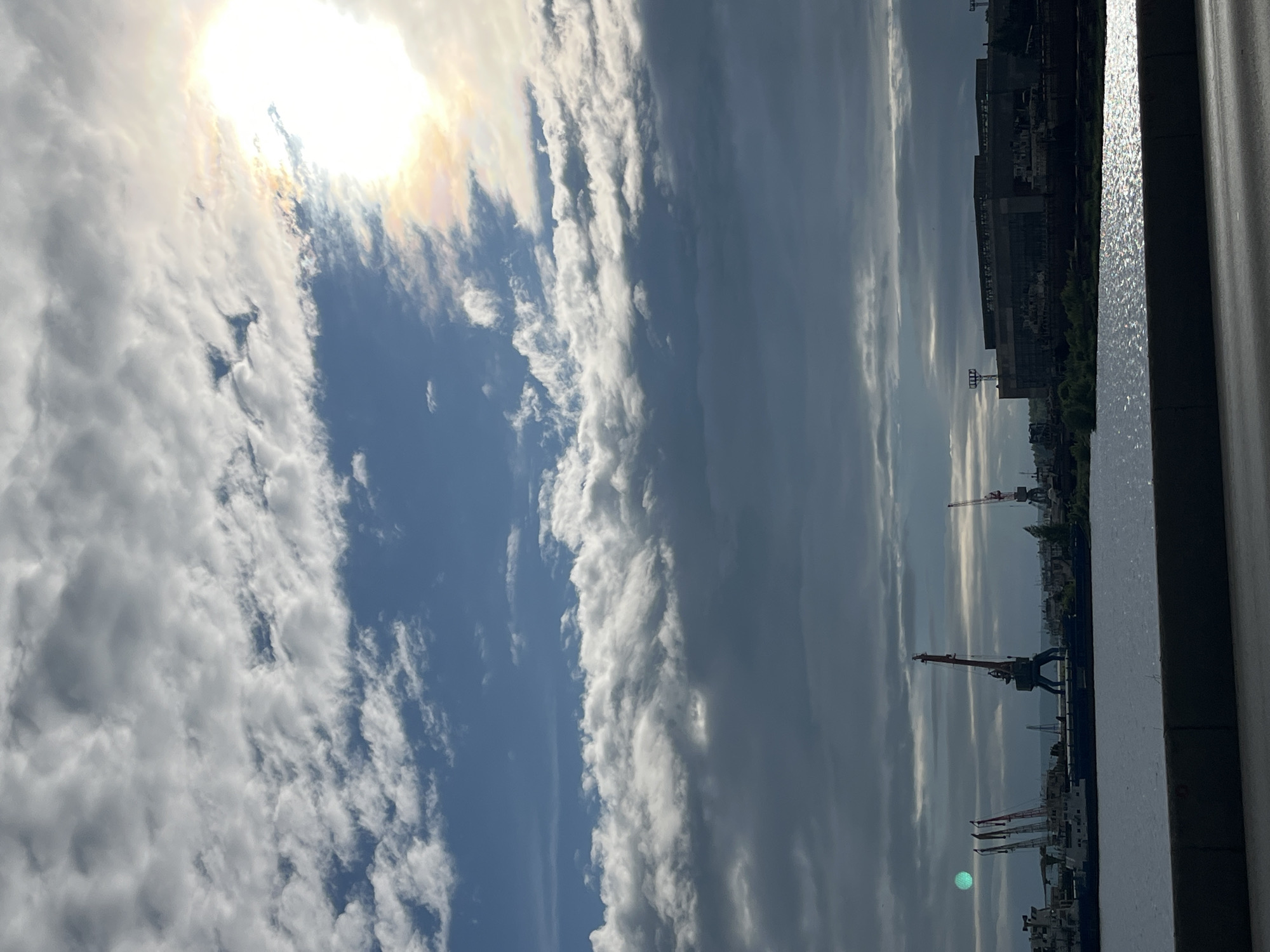 The image depicts a port scene in the city of Chaikovsky, Russia, on a partly cloudy day. The foreground shows a body of water with sunlight reflecting off its surface. In the mid-ground, a shoreline is visible with industrial structures, including ships and several large cranes. The sky is filled with a mix of white and dark grey clouds, with the sun partially obscured by clouds on the right side of the frame, creating a bright halo effect. A faint green circular object is visible in the sky on the left side of the image. The overall atmosphere suggests an active industrial area with dramatic cloud formations.