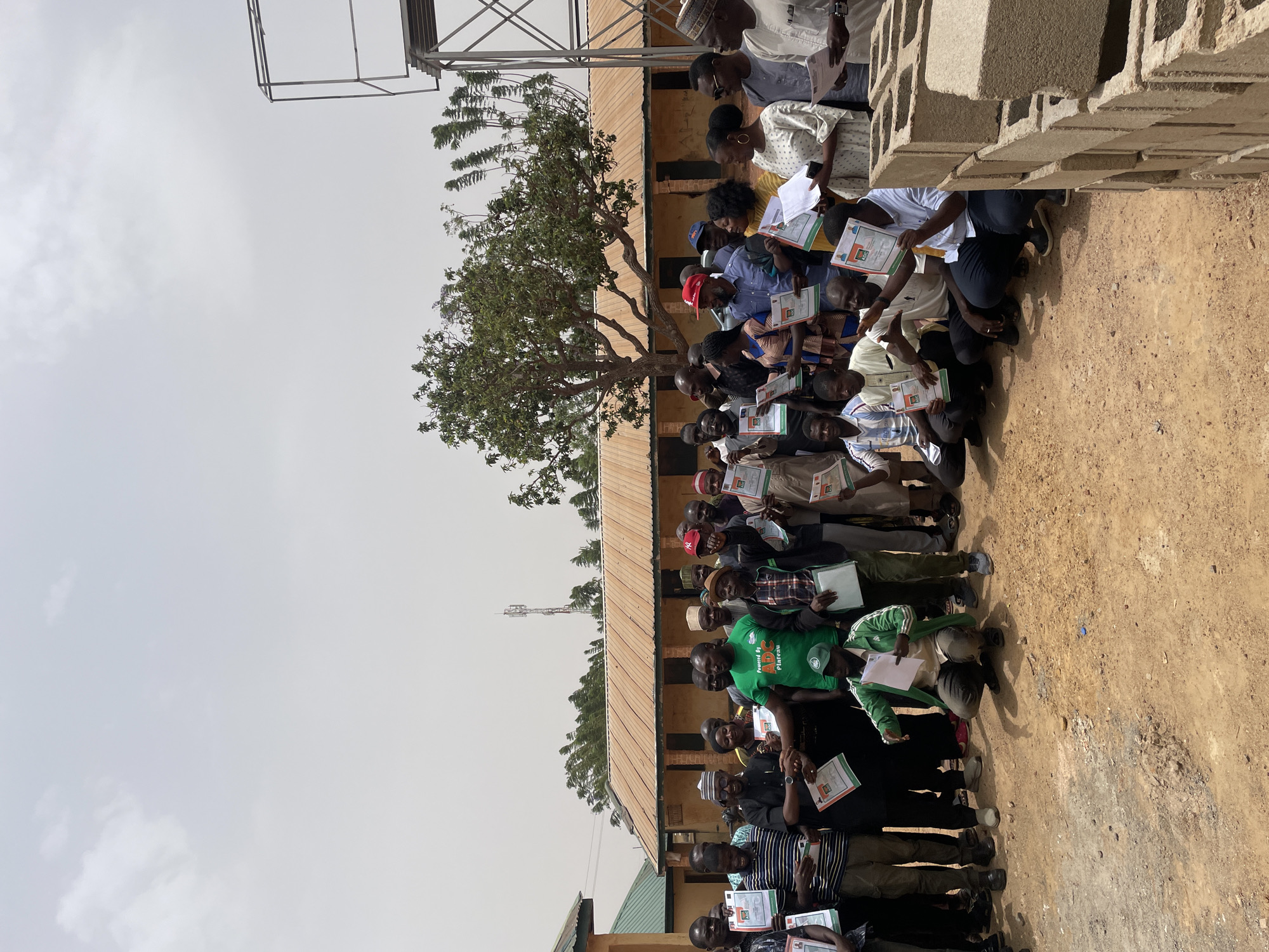 A daytime outdoor scene shows a group of approximately 40 people, mostly men, holding certificates or documents. They are standing close together in what appears to be a yard or open space with a dirt ground. The background includes a building with a rusty-colored roof, a tree, and a metal tower structure. A stack of concrete blocks are visible to the right. The sky is overcast. The location is likely in Jos, Nigeria, inferred from the style of dress and the context of the objects in the image.