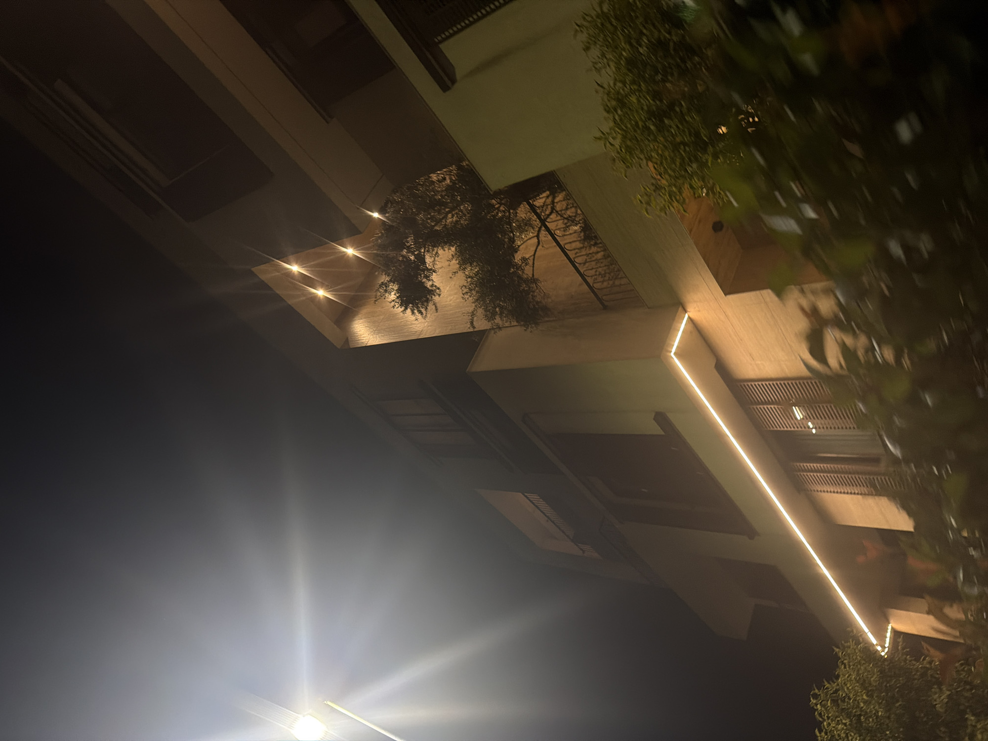 The media file displays a nighttime outdoor scene, captured with a rotated perspective, looking upwards towards a building facade and sky. The time of day is clearly night, characterized by a dark, almost black sky and prominent artificial lighting.
The central focus is a modern architectural structure, possibly a multi-story building, with clean lines and geometric shapes. Warm, artificial light emanates from several sources, creating bright starburst flares and illuminating portions of the building and surrounding elements. A prominent feature is a long, linear LED strip light running along an overhang or a façade element, casting a warm glow. Additionally, several recessed spotlights are visible on an architectural overhang in the upper-middle section of the image, illuminating what appears to be a balcony or a sheltered area with some visible foliage.
Dark green foliage, likely from trees or large bushes, is visible on the right side and growing from an upper level of the building, silhouetted against the illuminated structure and dark sky. The lighting highlights the texture of the building materials, which appear to be light-colored stone or concrete. The overall mood is quiet and atmospheric, typical of an evening architectural shot.
There are no people or specific activities taking place. No visible text is present in the image. The location is Podgorica, Montenegro.