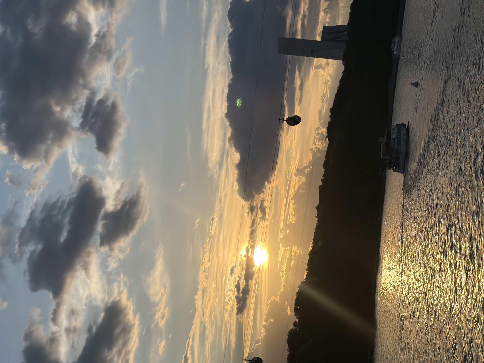 The image captures a picturesque sunset or sunrise over a wide body of water, likely the Moskva River in Moscow, Russia, as suggested by the location context. The sky is a prominent feature, displaying a mix of dramatic, dark grey cumulus clouds on the left and lighter, streaky clouds illuminated by the sun's warm, golden light towards the center and right.
The sun is a brilliant, bright orb positioned centrally, just above the horizon, casting intense golden rays across the scene. Its light creates a strong lens flare and reflects vibrantly on the textured surface of the water, forming a shimmering path of gold and orange that extends into the foreground.
A dark, undulating silhouette of land, densely covered with trees, stretches across the horizon, separating the sky from the river. Against the lighter sky behind this landmass, a tall, angular, light-colored building or tower is visible.
Several boats are present on the water: a larger, white passenger boat is seen on the right, and a smaller, less distinct vessel is further in the distance. Running horizontally across the sky, power lines or cables are visible, from which a dark, spherical cable car cabin hangs in the mid-ground, suggesting the presence of a cable car system like the Moscow Cable Car. Another similar cabin is faintly visible to the left.
A notable and unusual detail is a distinct, bright green, grid-like pattern appearing within a dark cloud in the upper-middle left section of the image. This is an optical artifact, likely a lens flare or internal reflection caused by the bright sun.
The overall atmosphere is serene and beautiful, characteristic of the golden hour, with no visible text in the image.