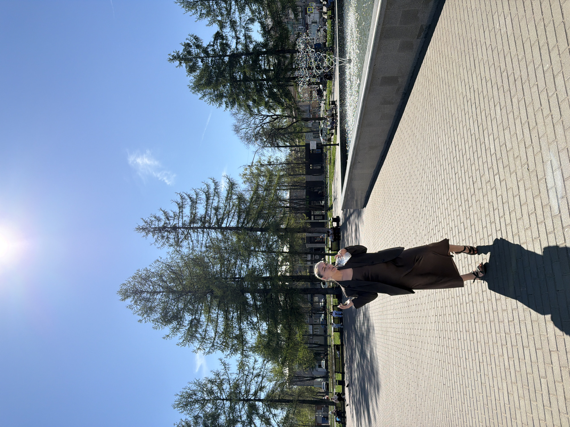 A woman stands on a paved area in Chaikovsky, Russia, holding a water bottle. She is dressed in a brown dress and a black jacket, wearing sandals. The background features tall trees, a fountain, and a clear blue sky with a visible contrail. Buildings are visible in the background, as are benches and people seated on the benches. The sun is shining brightly from the left side of the image.